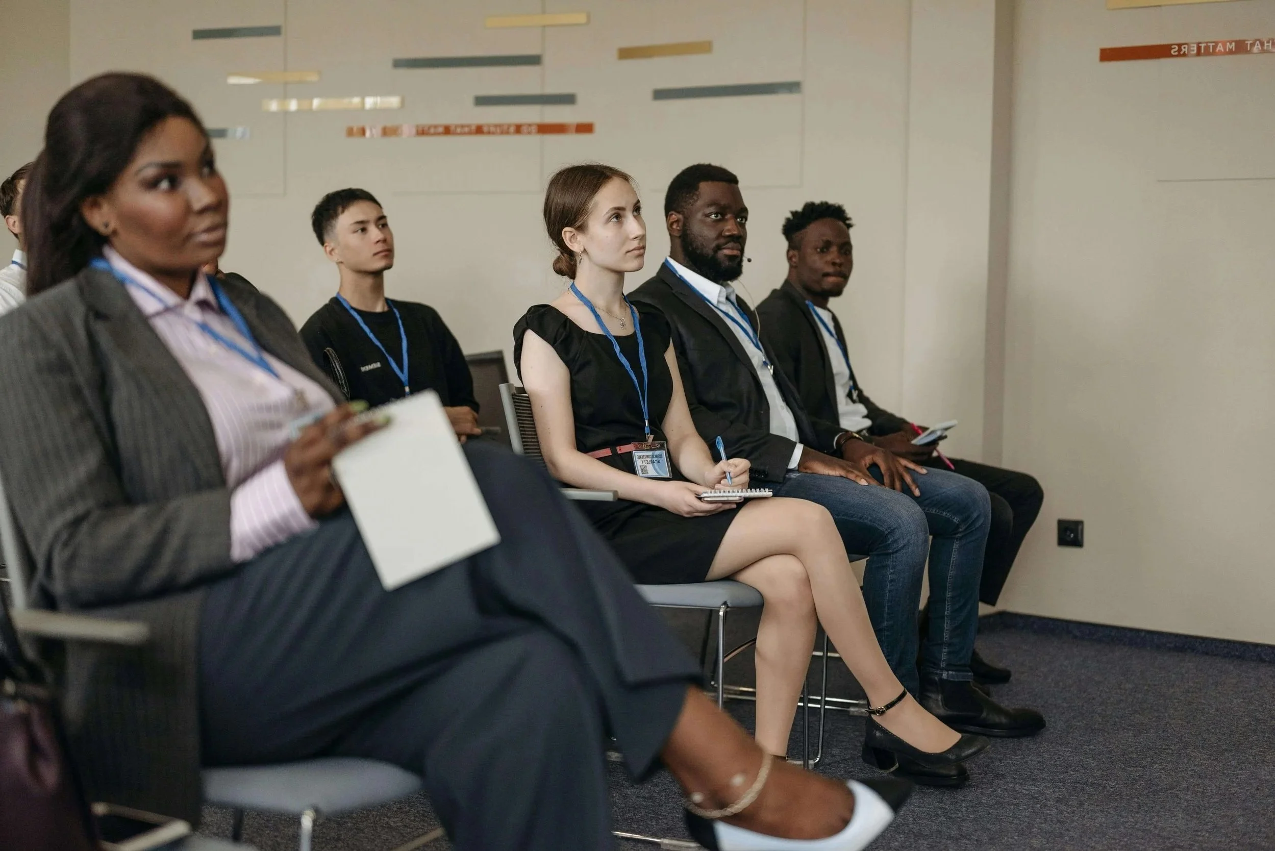 A group of diverse conference attendees sitting in a row, listening attentively, some taking notes on notepads and others holding smartphones.