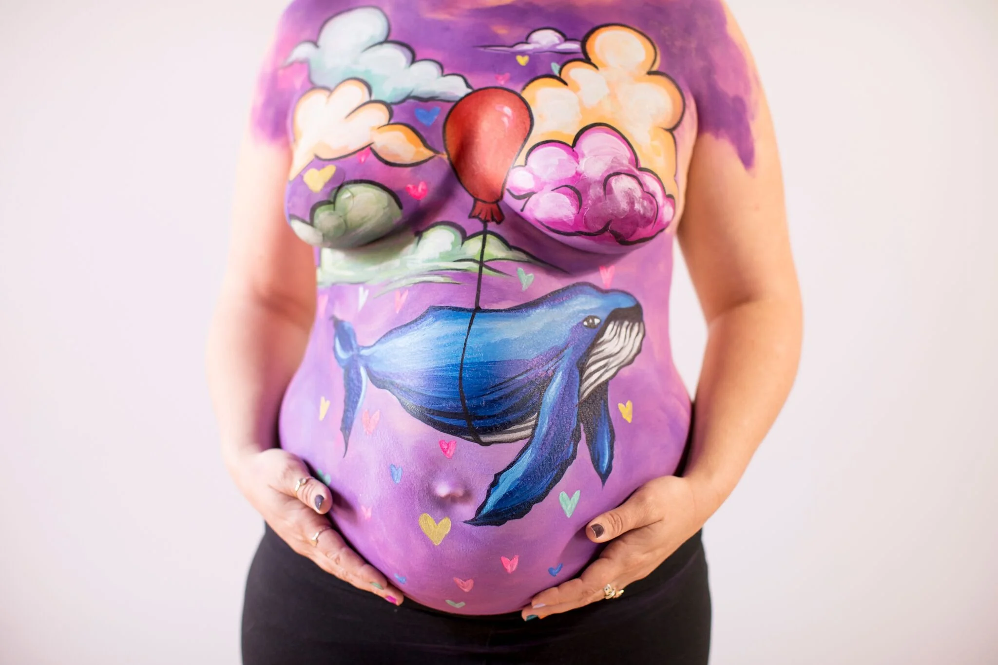 A pregnant woman with colorful body art depicting a whale swimming in water, surrounded by clouds, hearts, and a balloon shaped like a balloon in her painted stomach area.