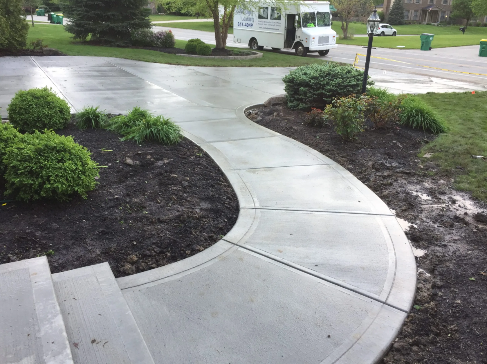 Freshly poured concrete walkway with landscaping and bushes, adjacent to a driveway. A white service truck is parked on the street.