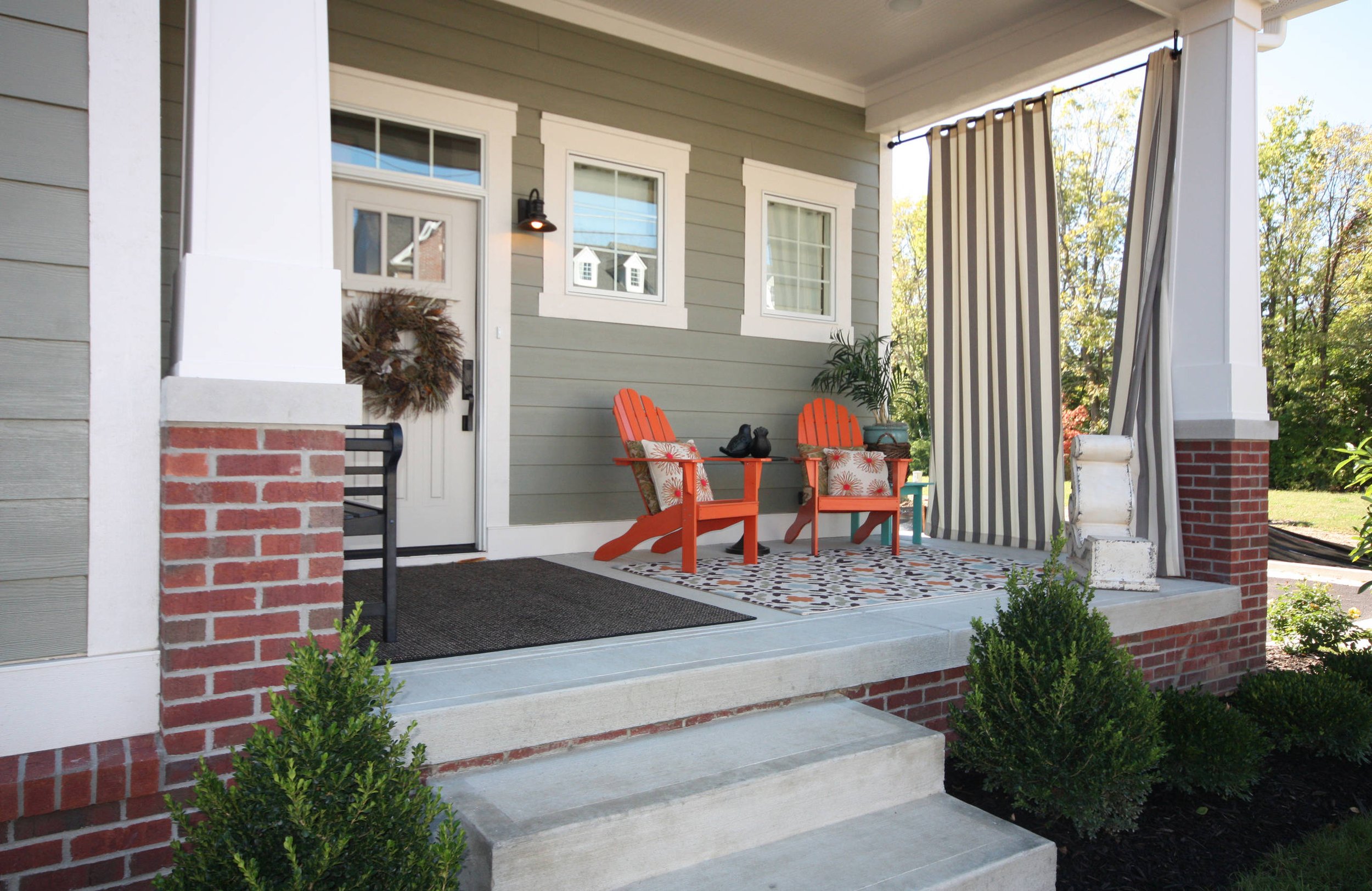 Front porch with two orange Adirondack chairs and a small table, decorated with patterned pillows. A door with a wreath, striped curtains, potted plants, and a patterned rug complete the setup.