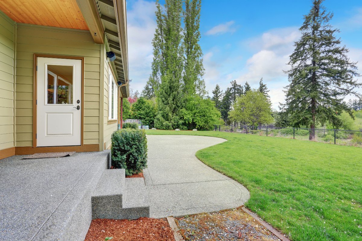 Backyard view with a concrete patio, steps, and grass lawn, next to a tan house, surrounded by trees and a fence.