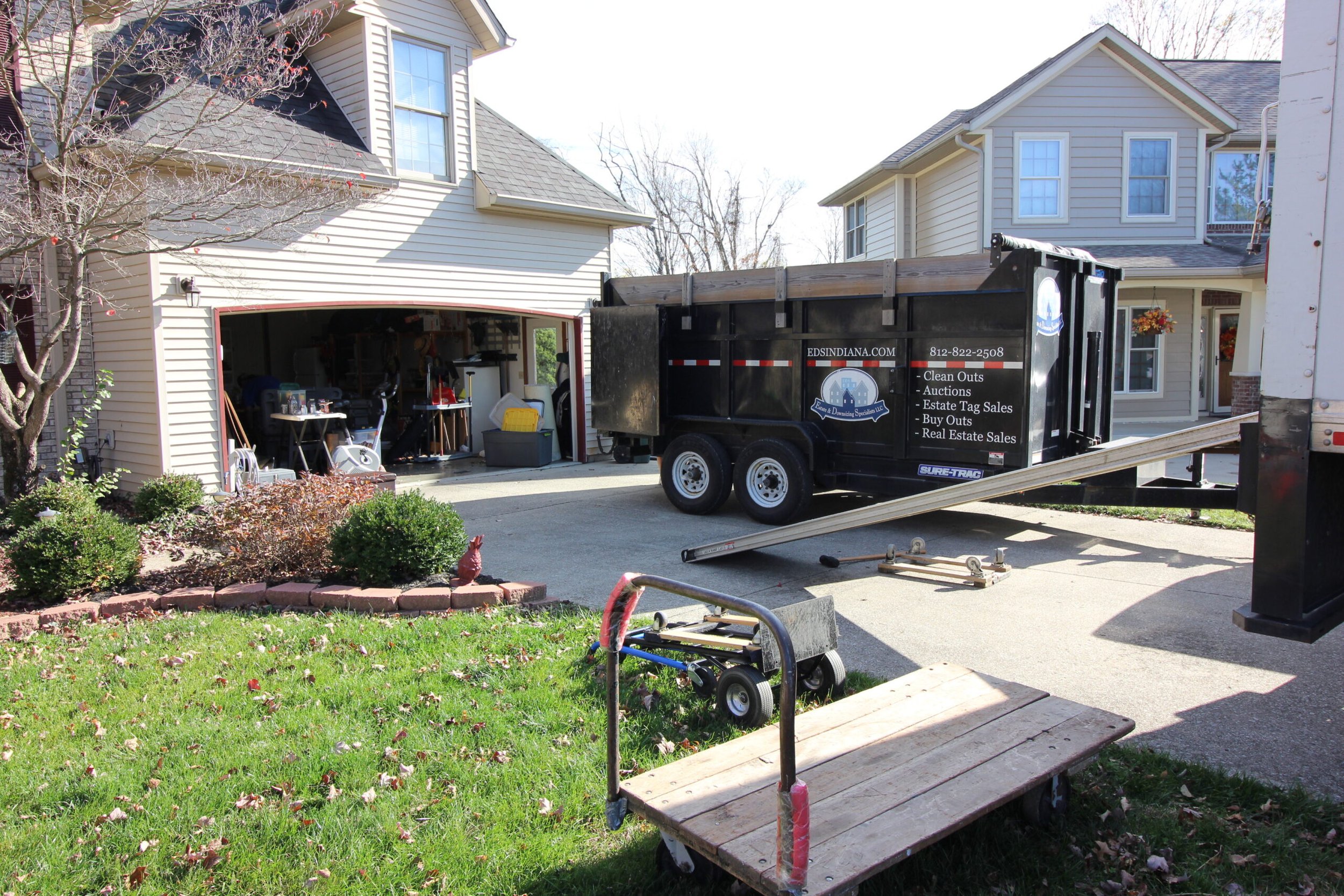 Residential garage with open door revealing clutter, moving dolly, and a large trailer parked in driveway with signage for clean outs and estate sales.