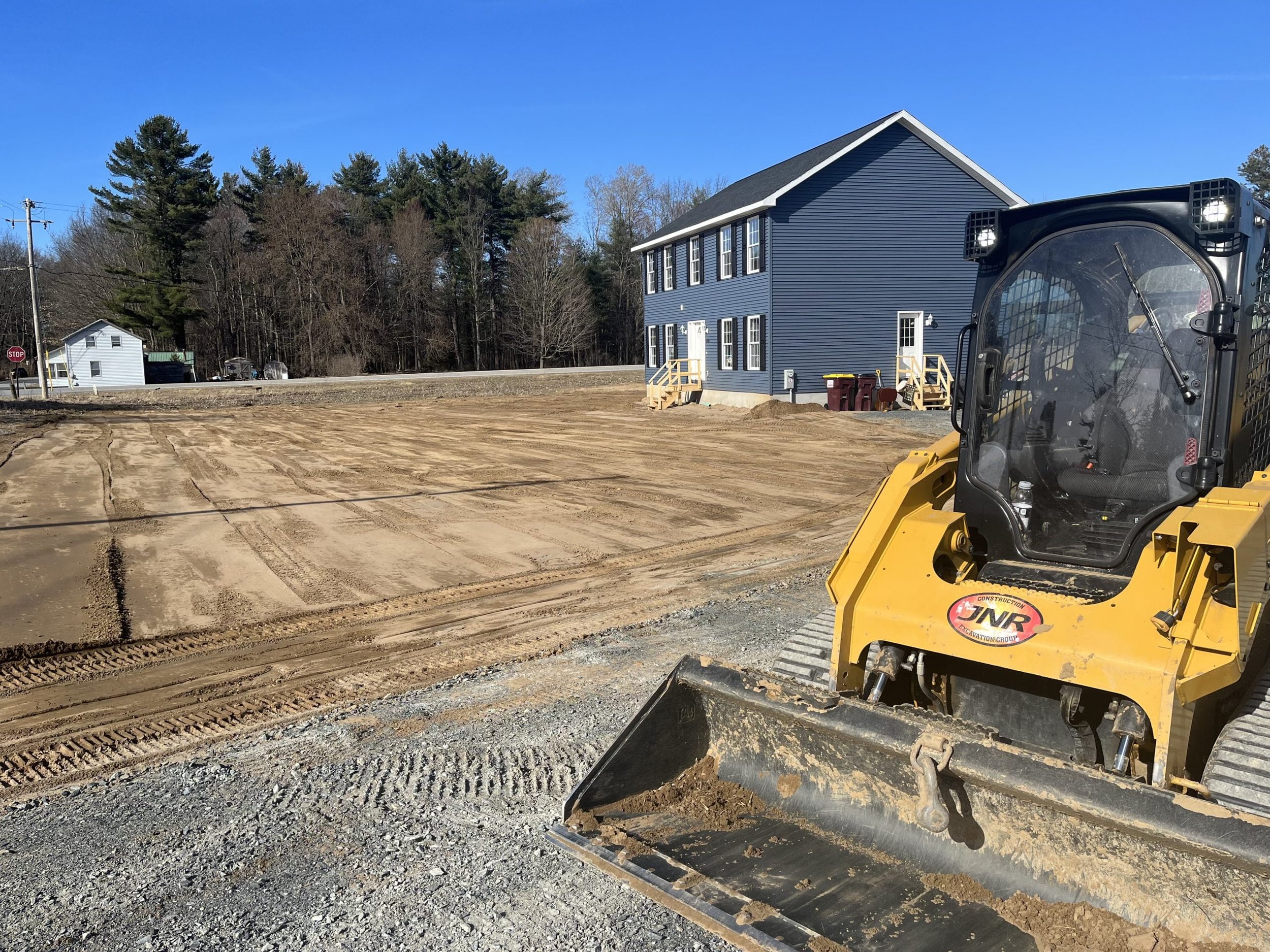 Construction site with a yellow skid-steer loader on a dirt lot in front of a blue two-story building, surrounded by trees and a clear sky.