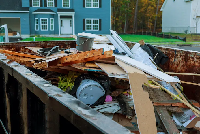 A dumpster filled with construction debris including wood, metal, and plastic, in front of a residential house.