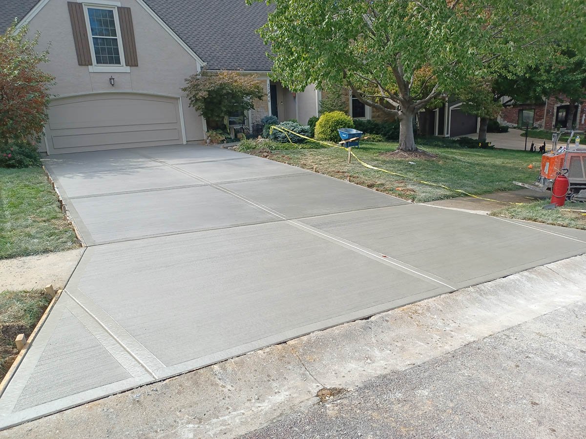 Newly poured concrete driveway in front of a house, with a garage and surrounding greenery.