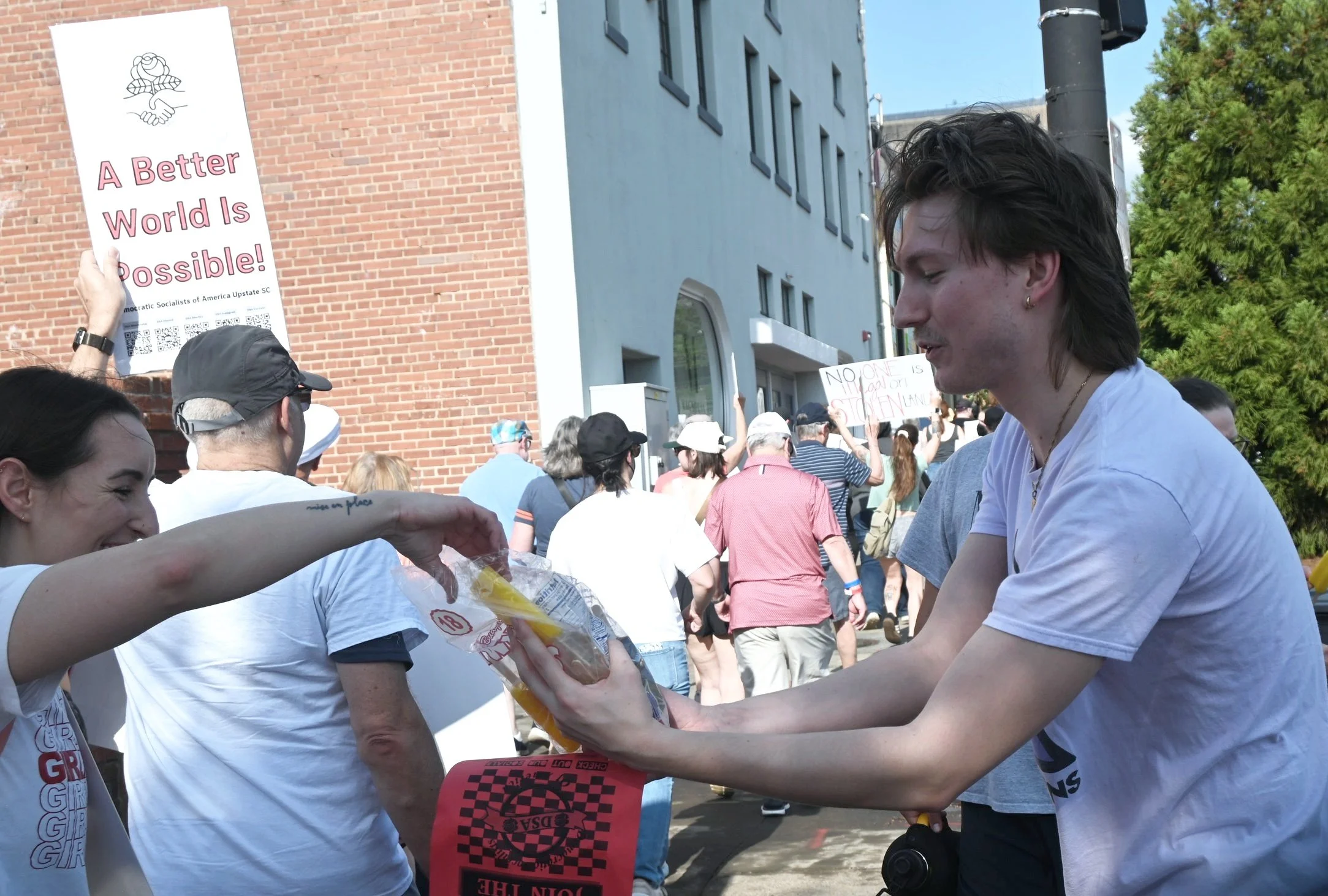 People participating in a street protest or rally, holding signs with messages advocating for a better world, with a brick building and trees in the background.