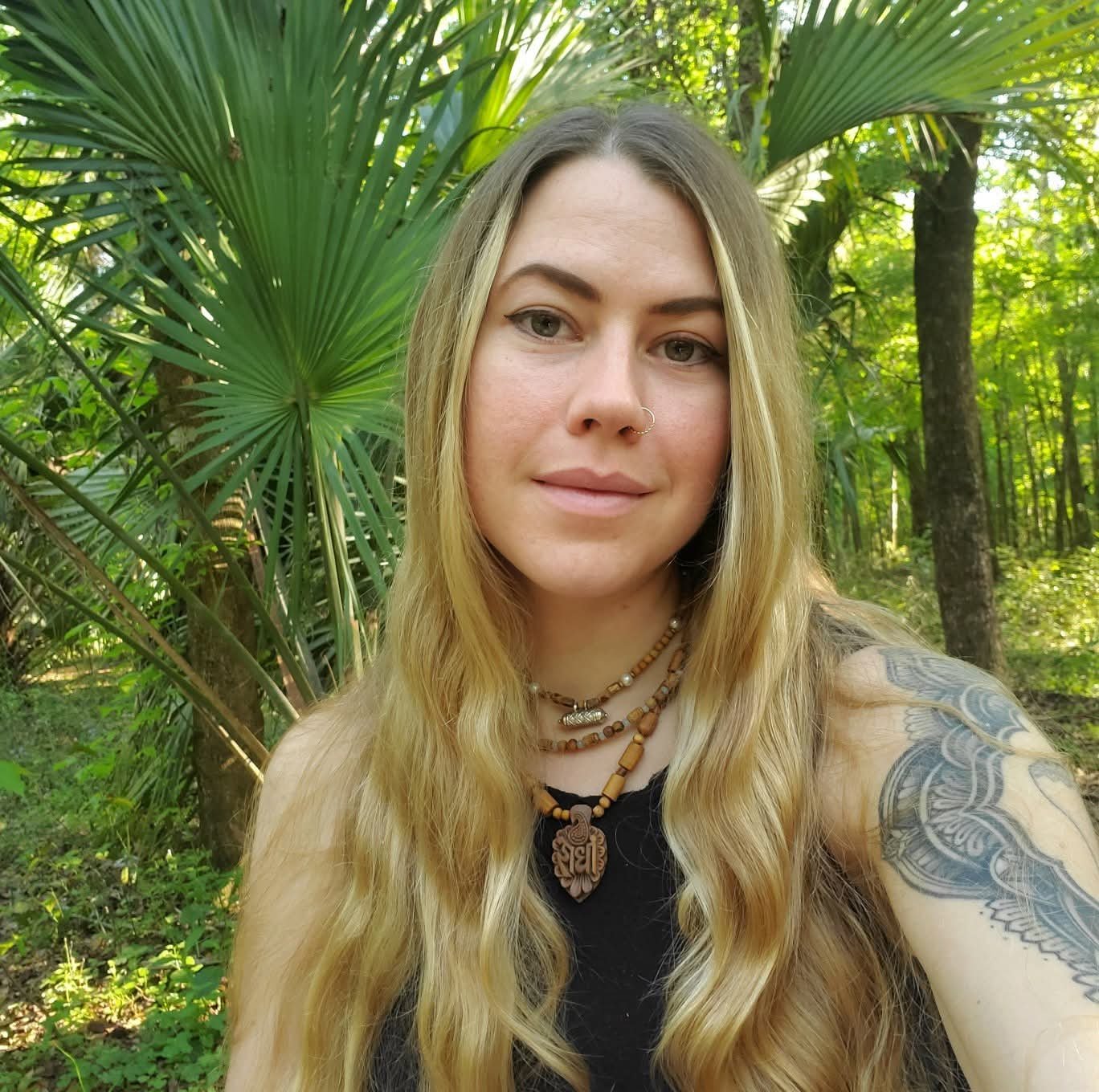Photo of Corinna Roberge in a forested place. She is smiling gently and wearing a black shirt and a wooden pendant
