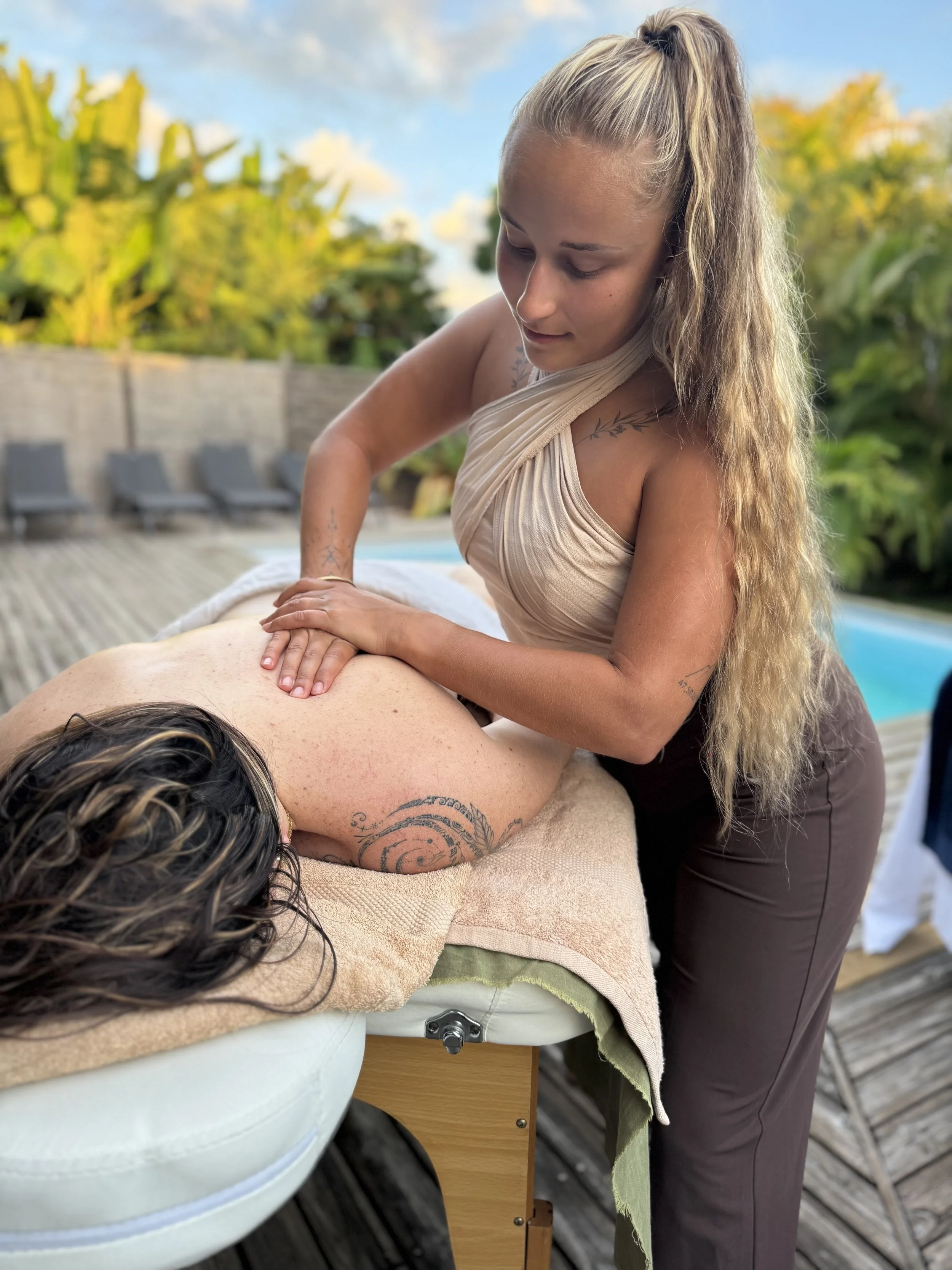 Une femme donne un massage à une personne allongée sur une table en extérieur, entourée de verdure et de chaises longues.