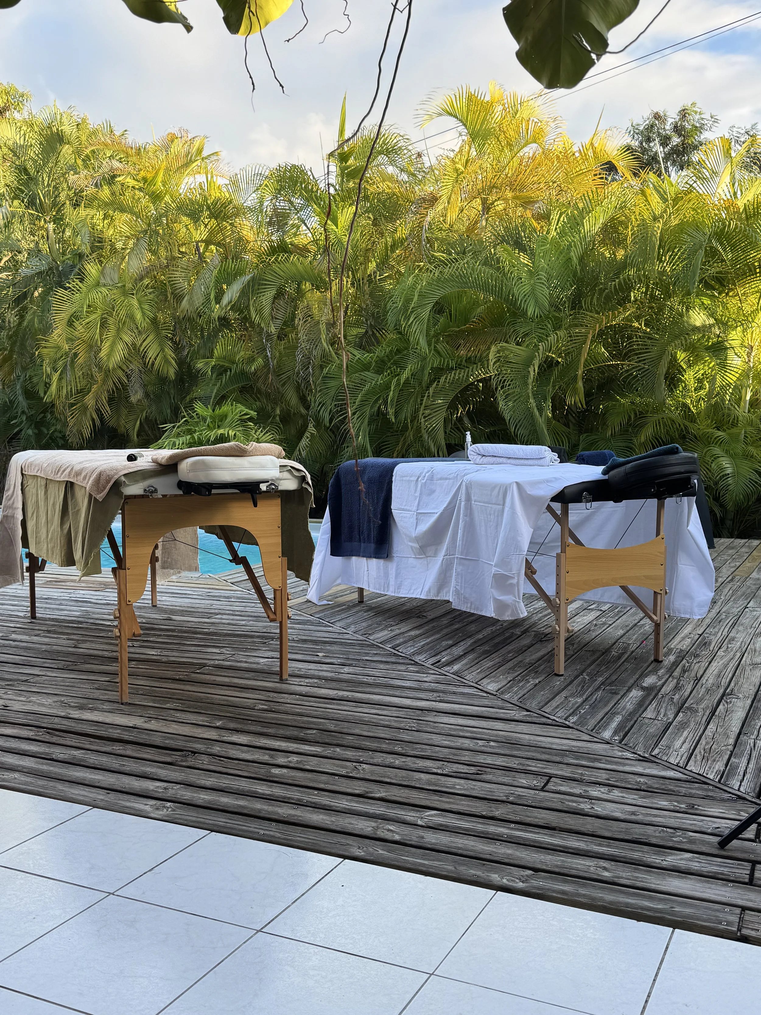 Table de massage avec des serviettes et des instruments de massage, située sur une terrasse en bois, avec un mur végétal dense de plantes tropicales en arrière-plan.