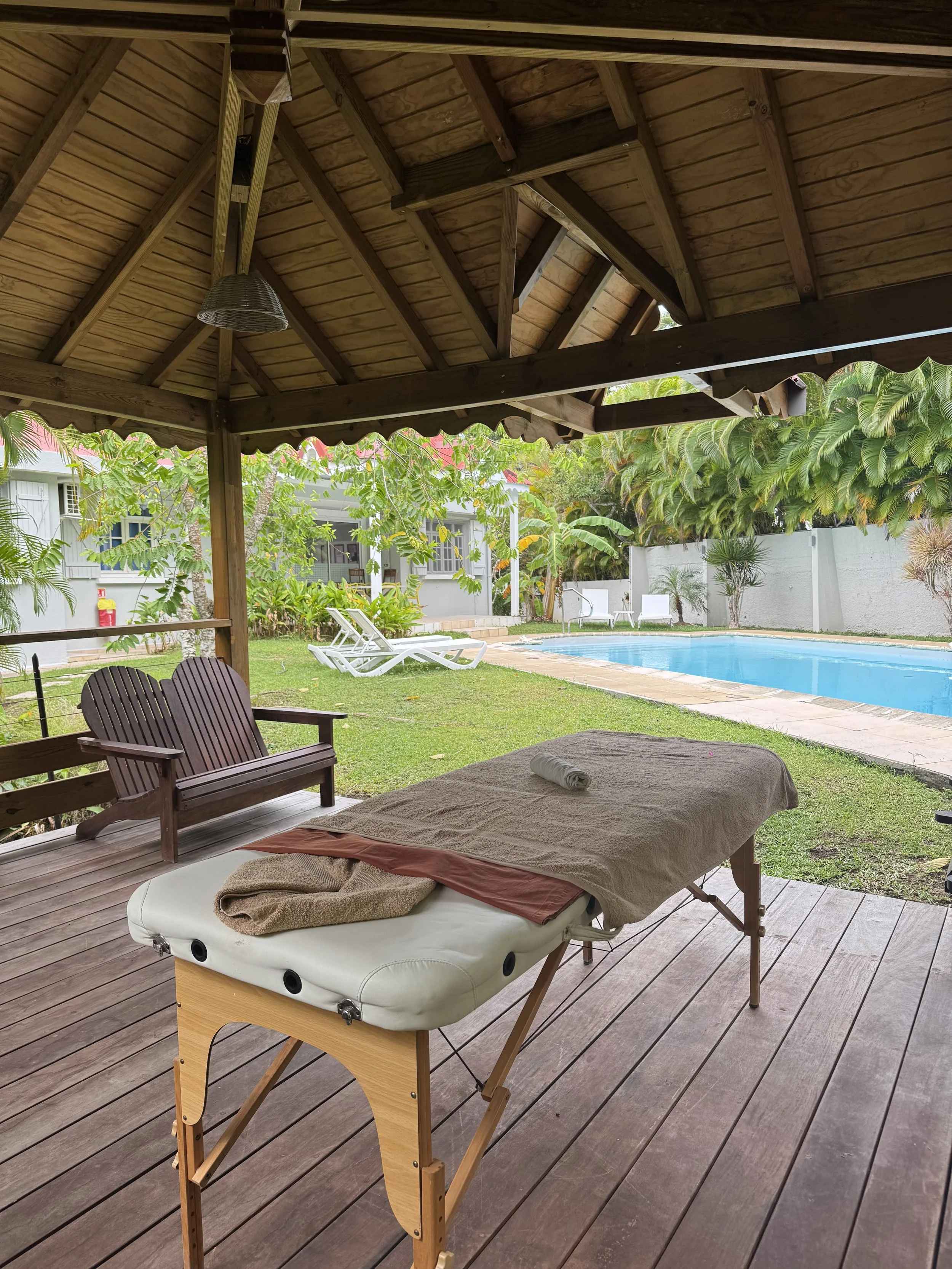 Une terrasse en bois couverte avec un lit de massage, une chaise en bois et une vue sur la piscine et le jardin tropical.
