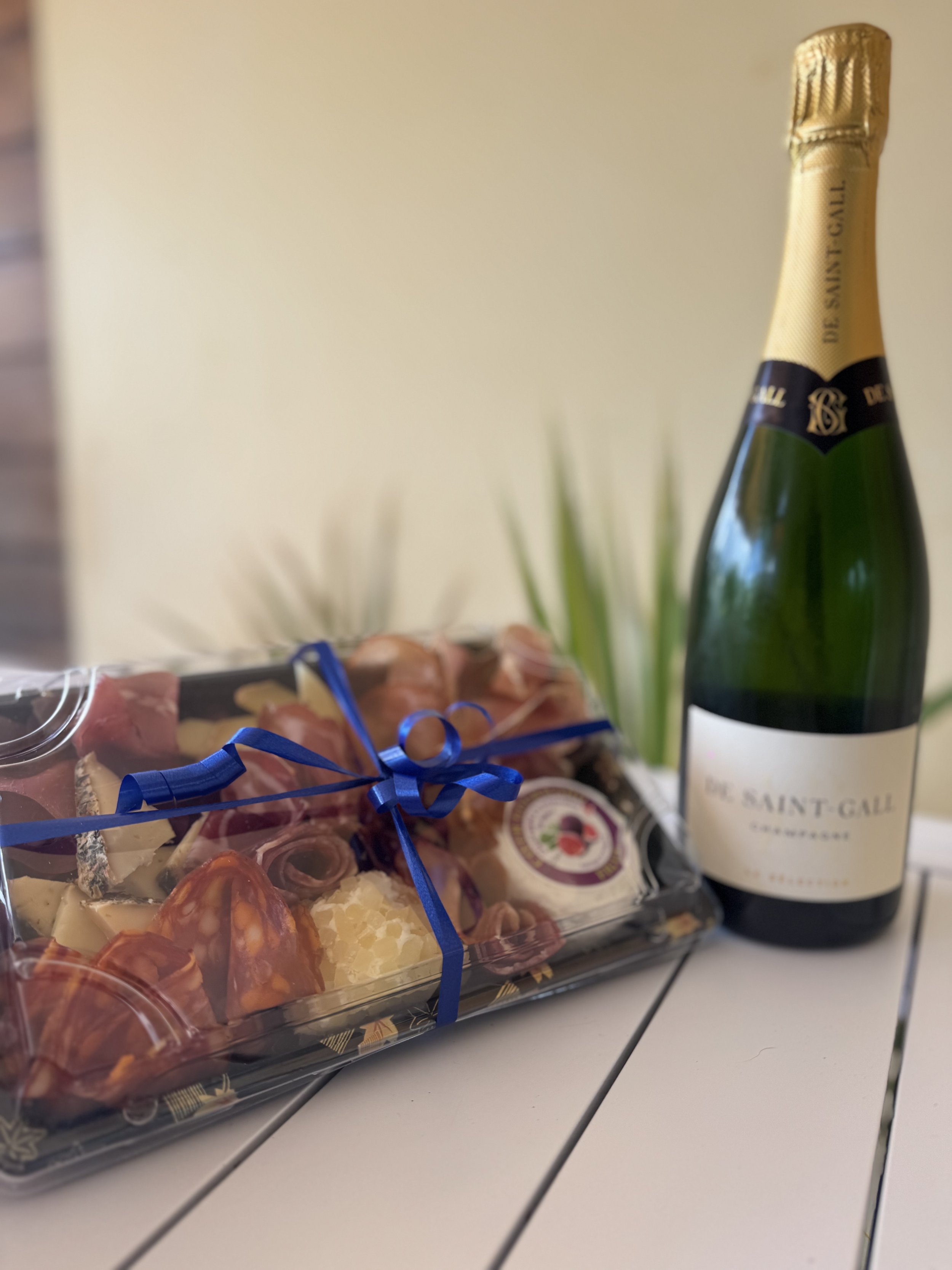 Une bouteille de champagne et un plateau de charcuterie avec des tranches de viande, du fromage, et des tomates cerises, décoré avec un ruban bleu.