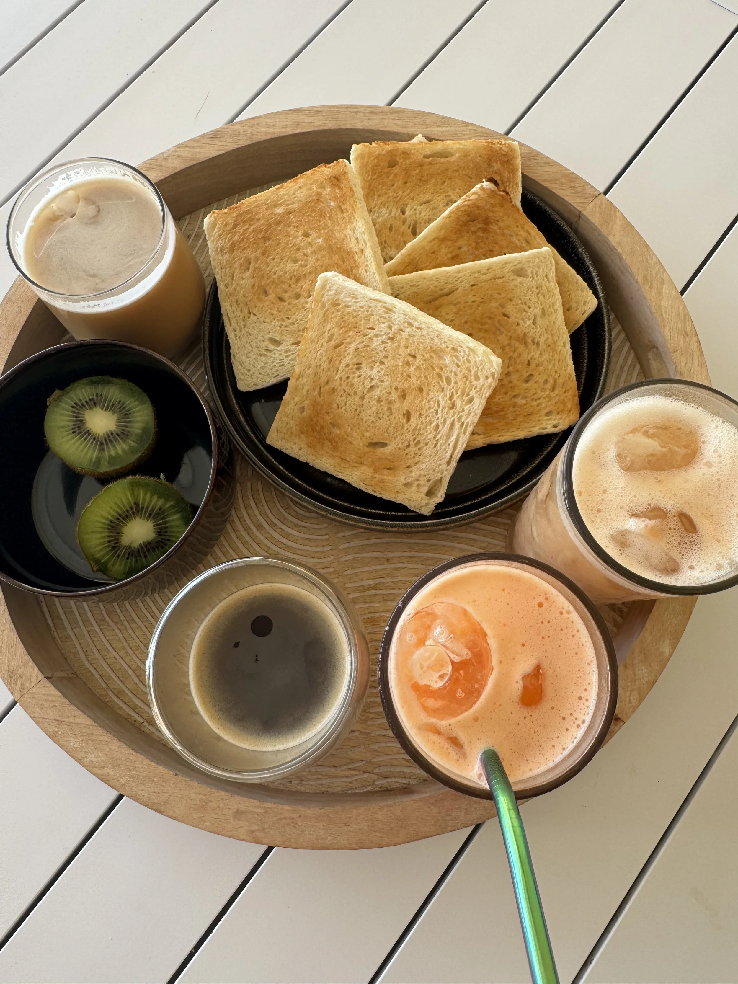 Un plateau en bois avec des tranches de pain grillé, deux verres de boissons colorées avec des glaçons, et une petite coupe avec des tranches de kiwi. La table est blanche à rayures.