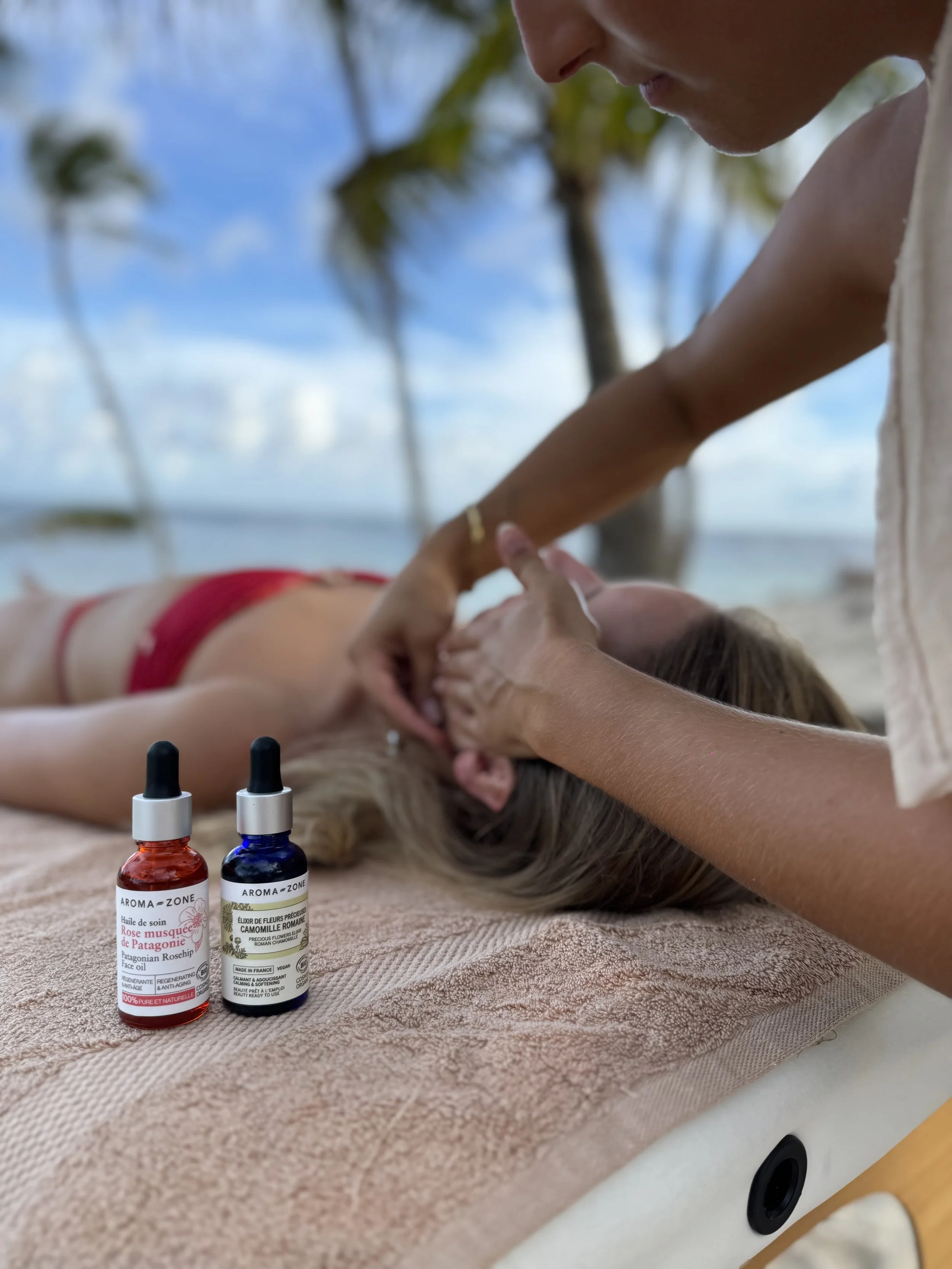 Une personne reçoit un massage sur un lit de plage. Deux flacons de produits aromatiques sont visibles à l'avant sur une serviette beige.