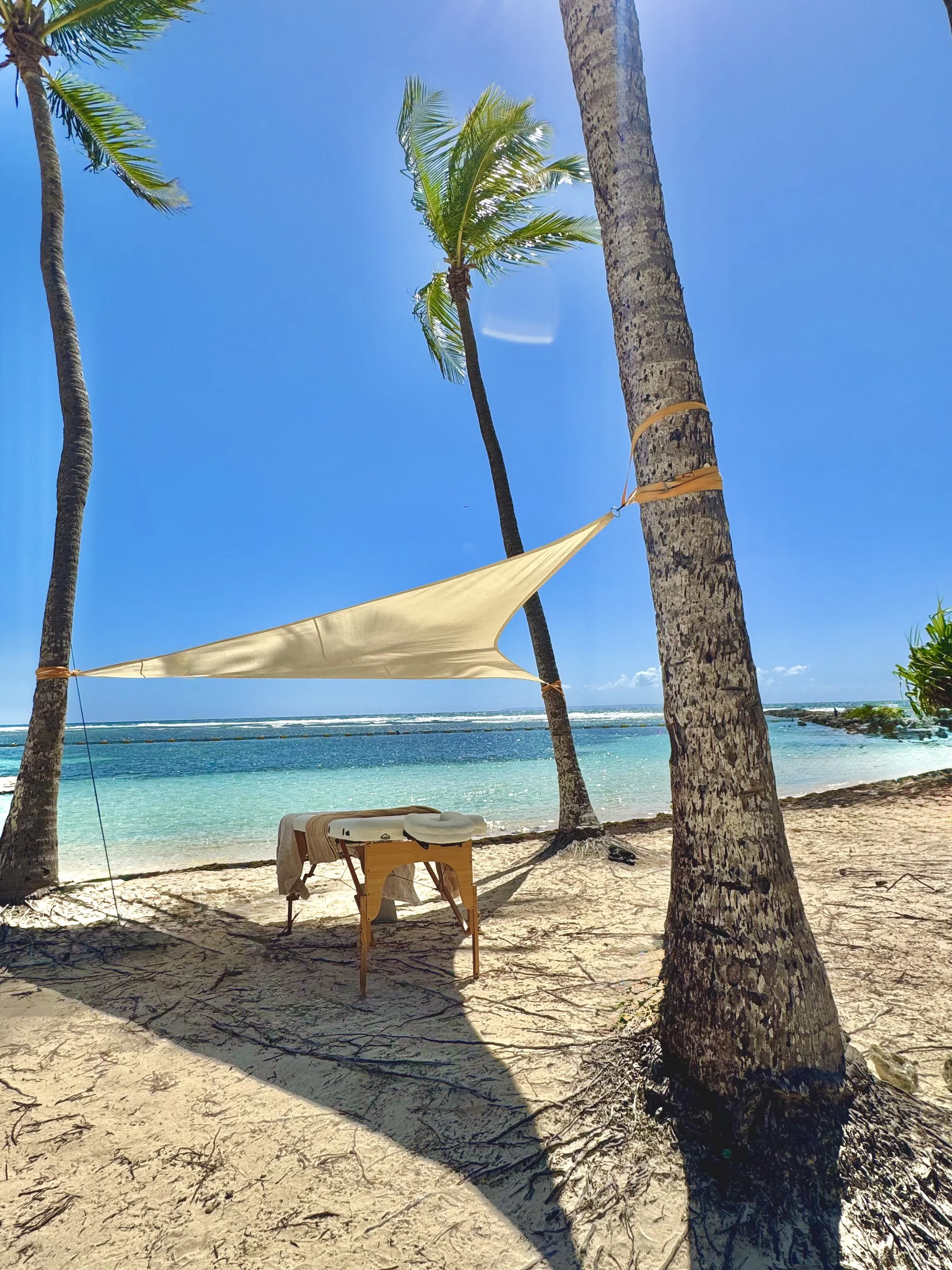 Plage de sable blanc avec des palmiers et une voile de tente beige, vue sur la mer turquoise sous un ciel bleu clair, ambiance relaxante et ensoleillée