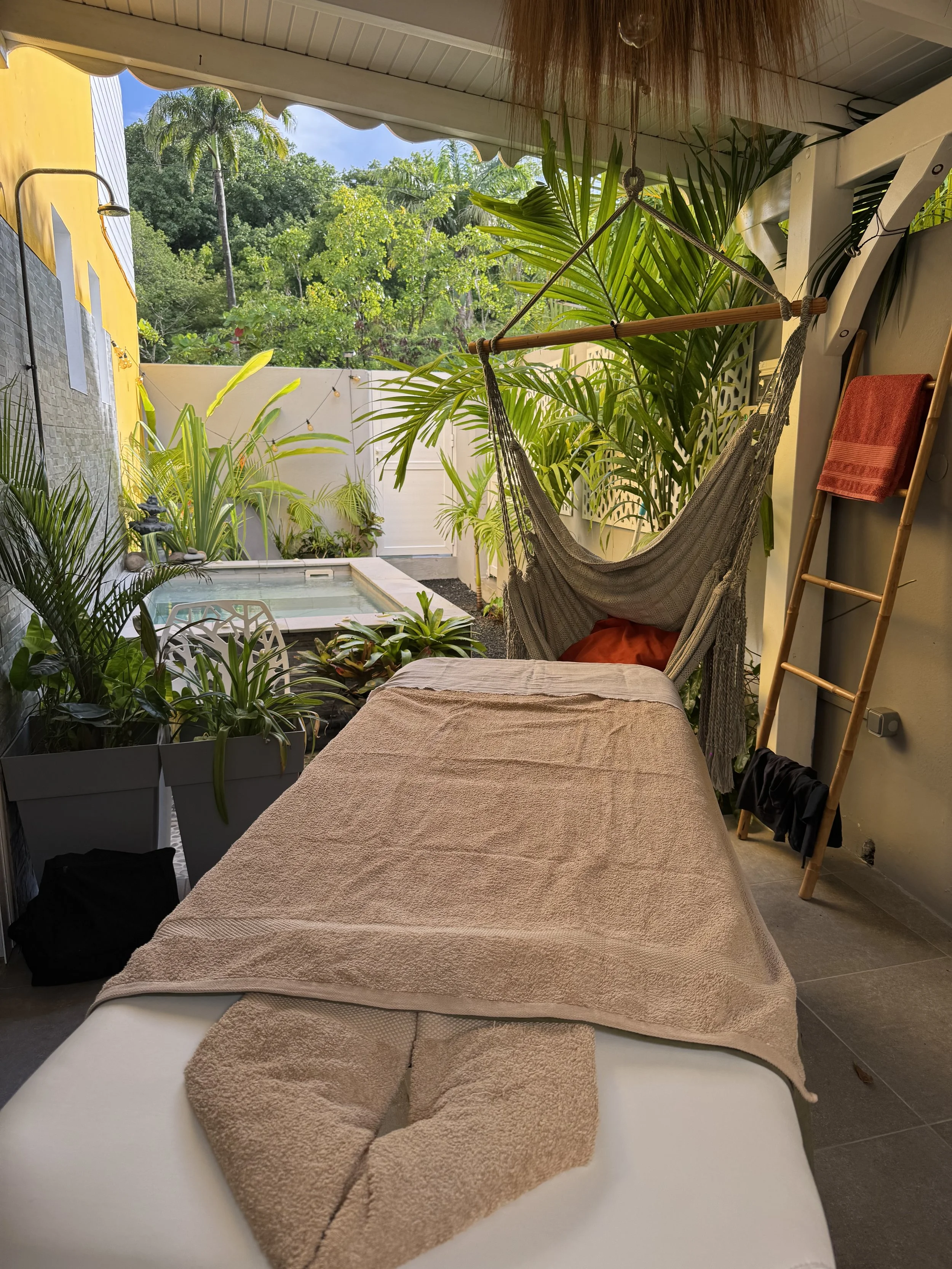 Zone de massage ou de spa extérieure avec un lit de massage recouvert d'une serviette beige, plusieurs plantes tropicales, un hamac suspendu, un châle ou un tapis rouge sur une chaise en bois, une douche en extérieur, et un bassin d'eau ou un spa, en