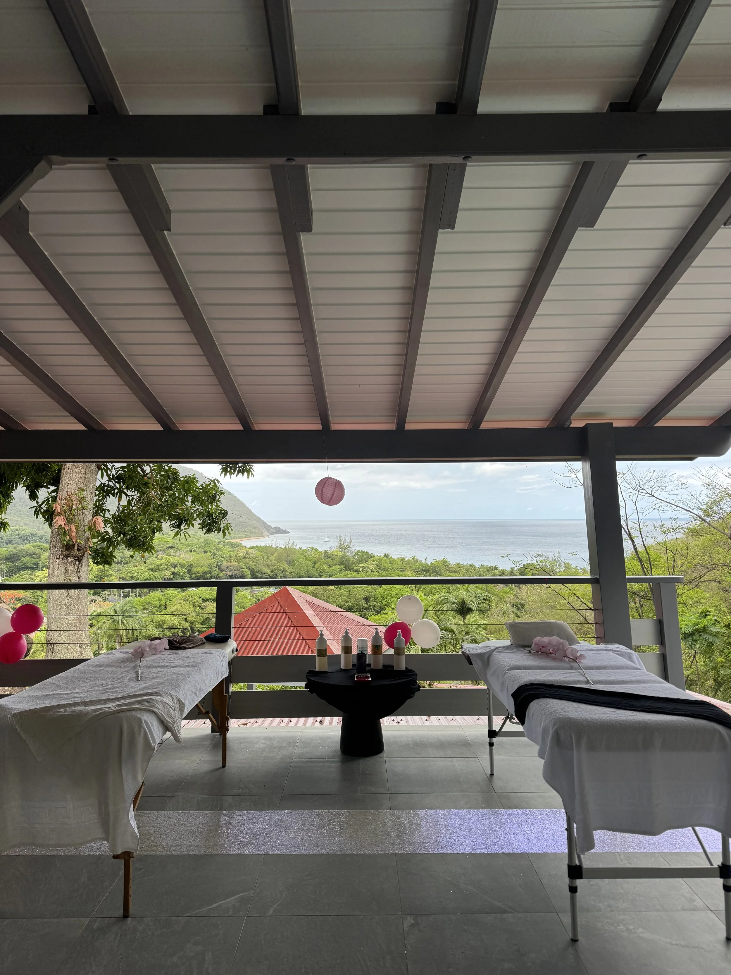 Deux tables de massage avec des draps blancs, décorées de ballons roses et blancs, dans une salle avec vue sur la mer et la nature, avec un toit en bois.