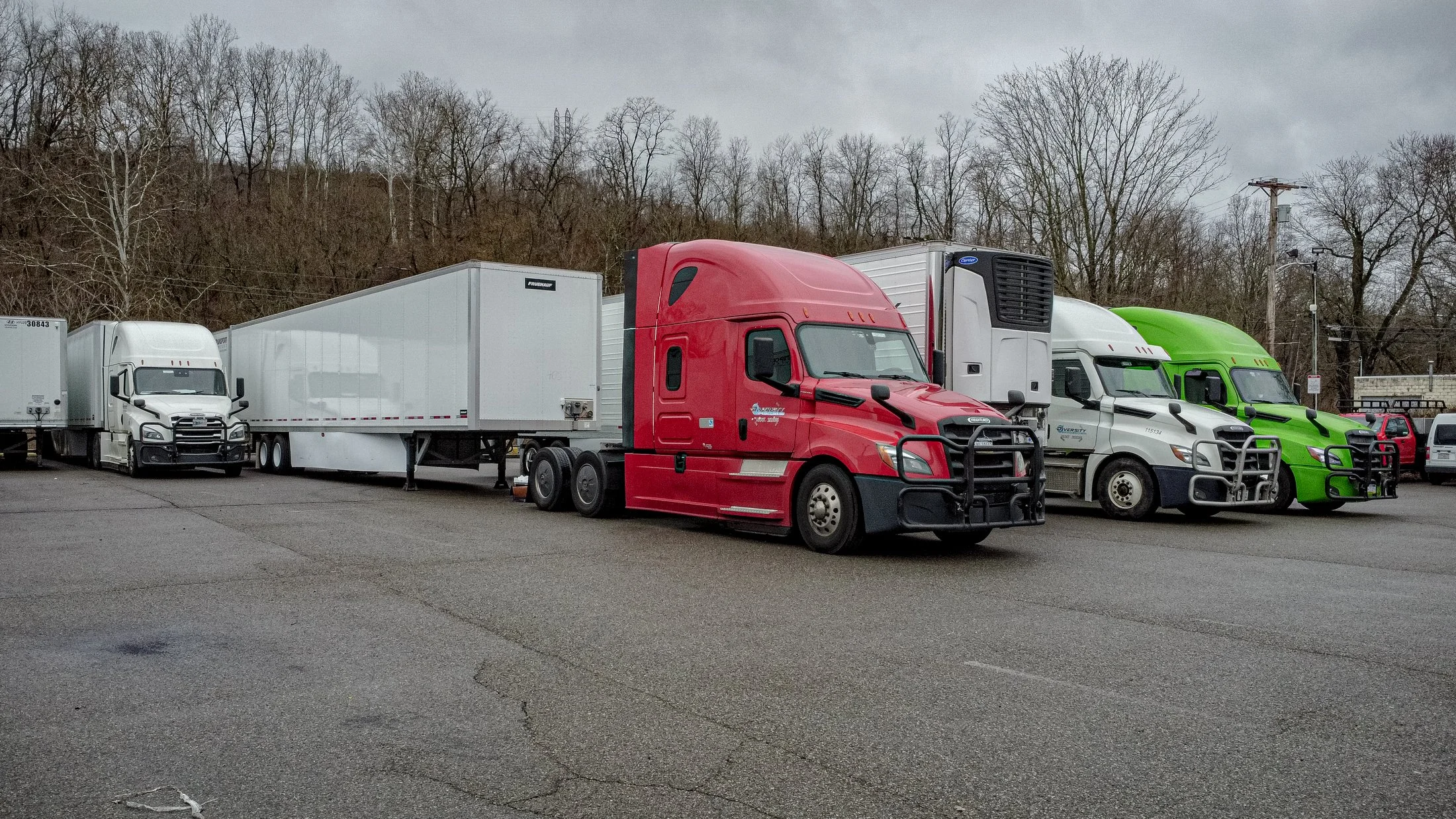 Parked semi-trailer trucks in a lot with bare trees in the background.