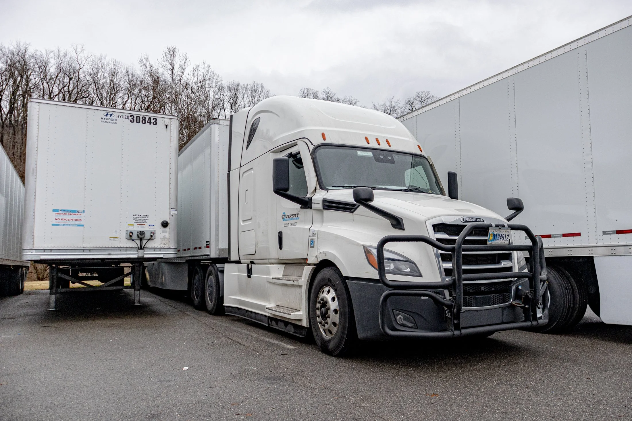 White semi-truck parked between two trailers in a parking lot, with trees in the background.