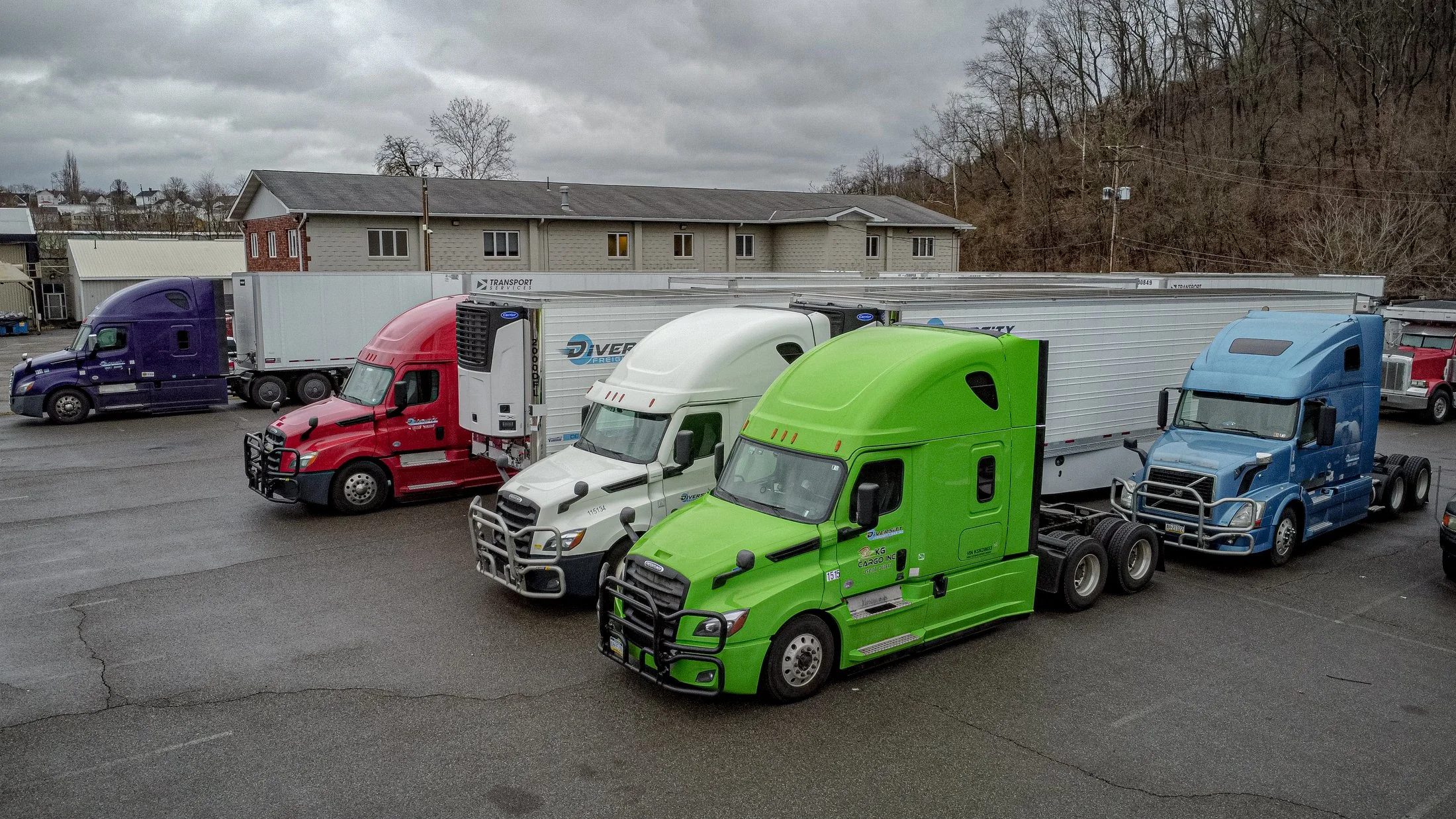 Several colorful semi-trucks parked in a lot with trailers attached, against a backdrop of buildings and a hillside.