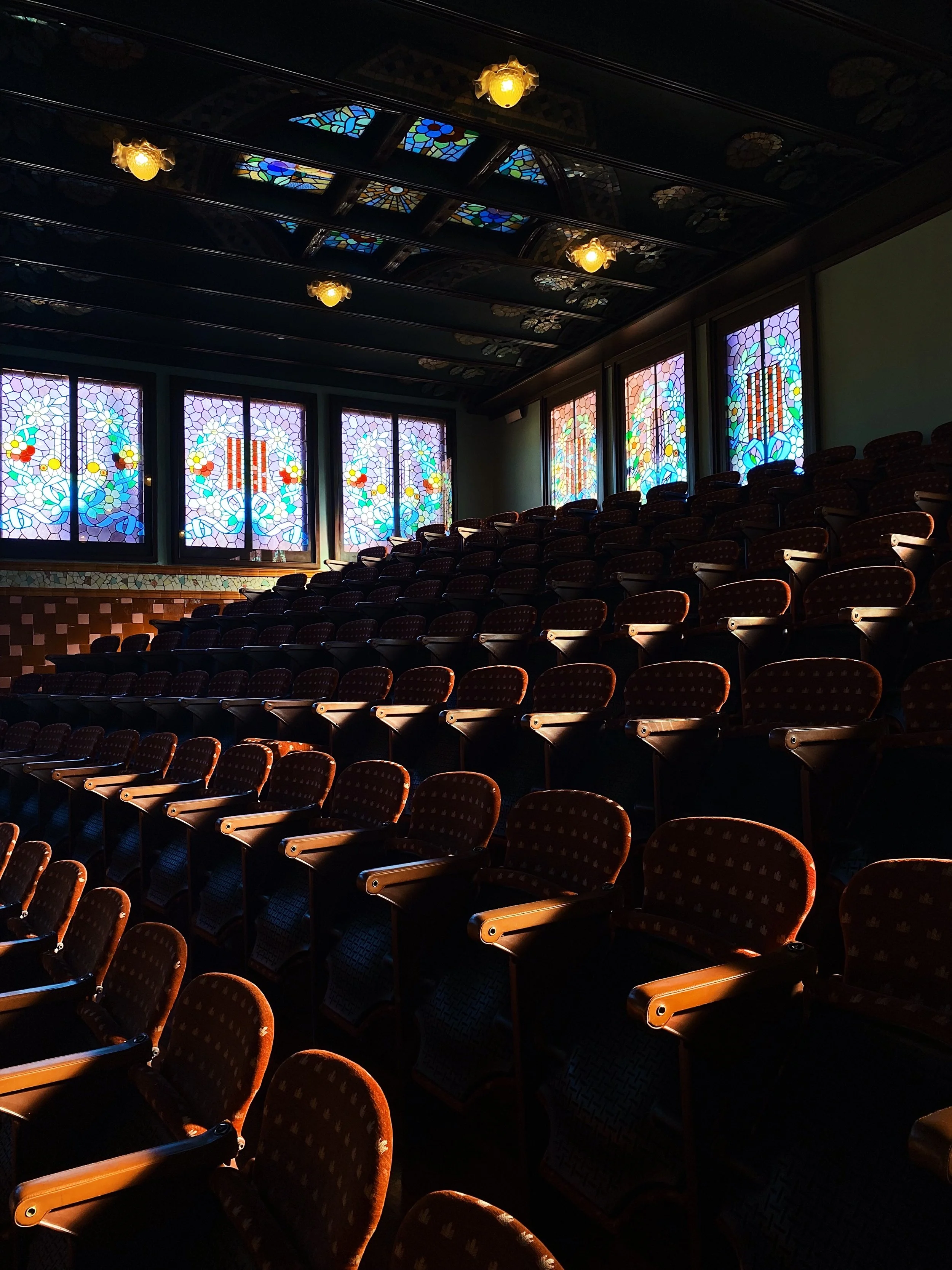 Empty theater with rows of red velvet seats, stained glass windows, and decorative ceiling with hanging lights.