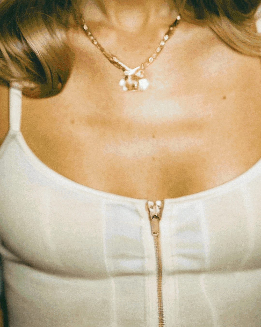 Close-up of a woman's upper chest and neckline, featuring a pearl necklace with a pendant and a white top with a zipper