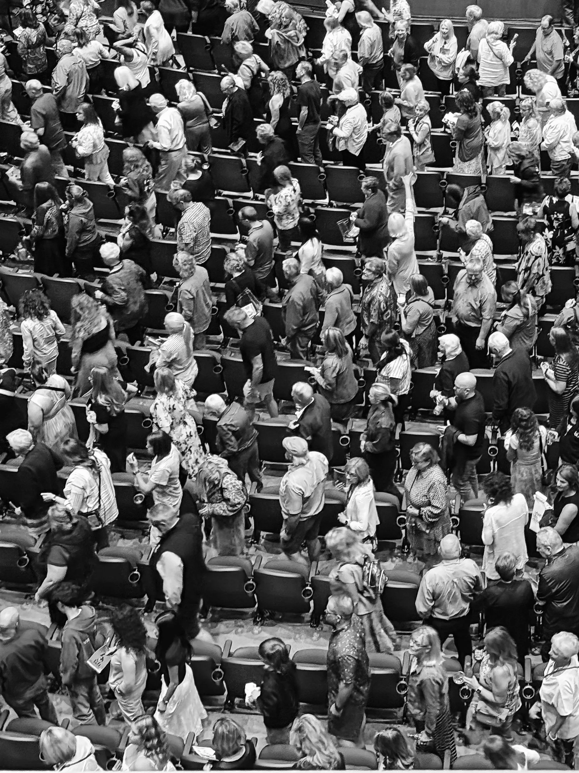 Black and white photo of a large crowd of people standing in an auditorium or conference hall, some holding papers or devices, arranged in rows of chairs.