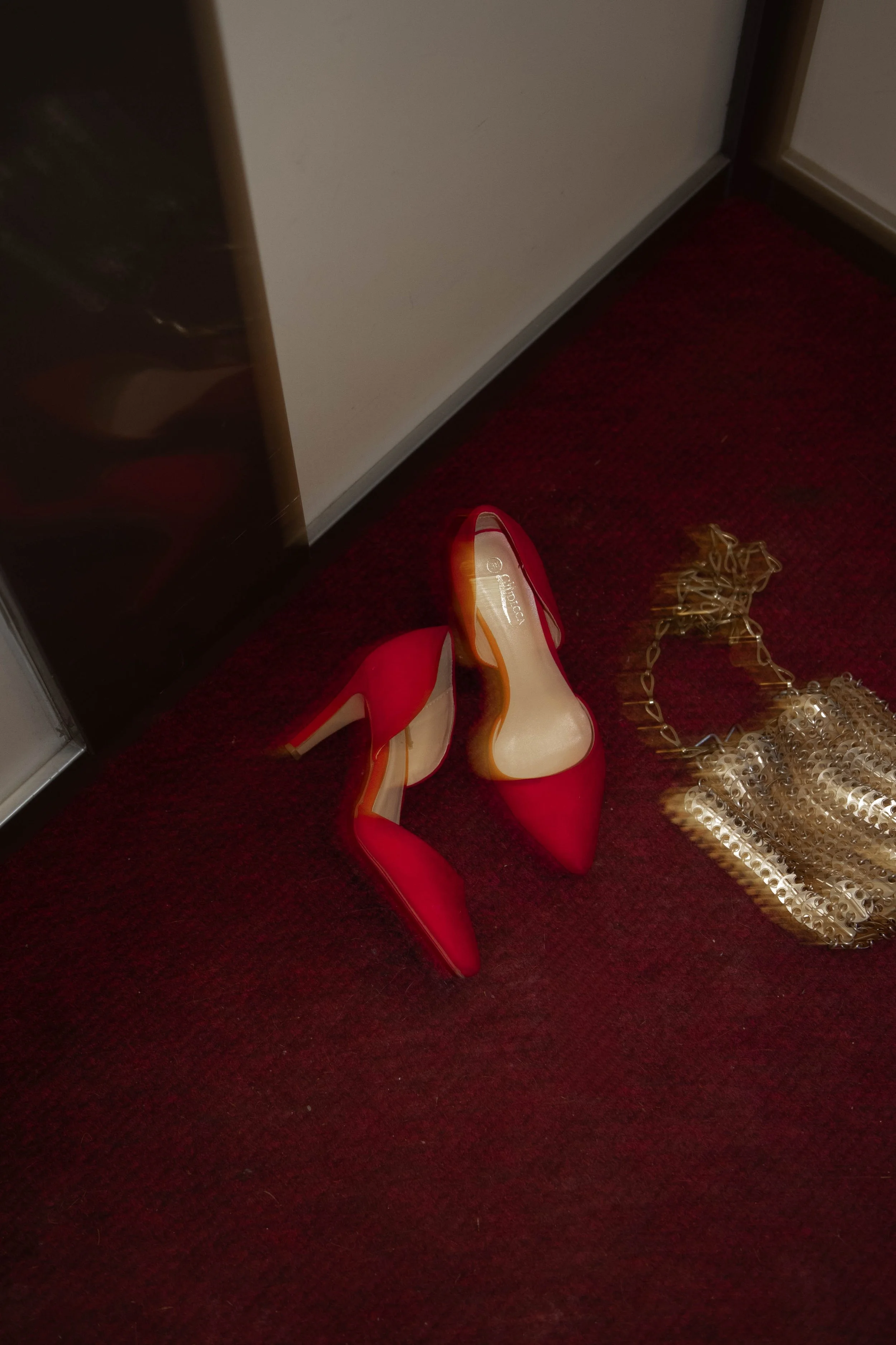 A pair of red high-heeled shoes on a red carpeted floor next to a gold-colored chain purse.