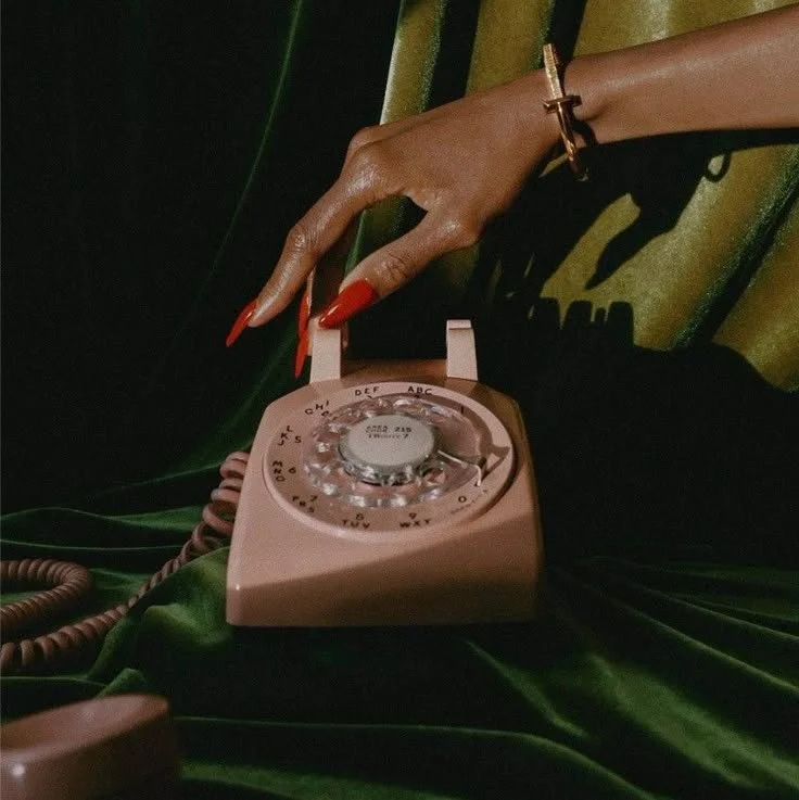 A hand with red nails and a gold bracelet reaching to adjust the dial of a vintage pink rotary telephone on green velvet fabric.