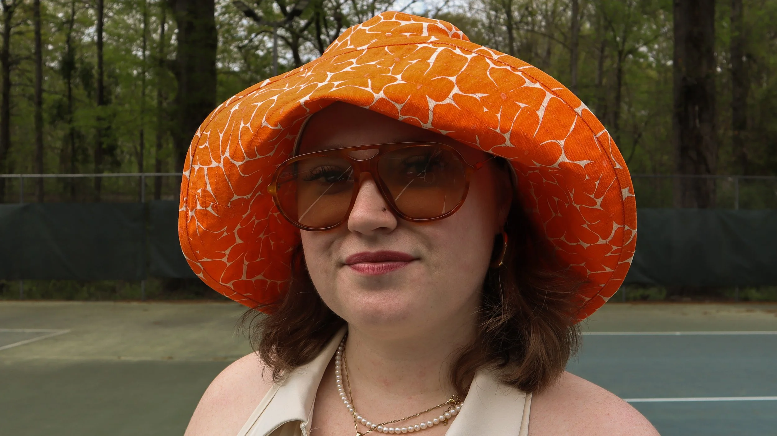 A woman wearing a large orange hat with a white pattern, brown sunglasses, gold hoop earrings, a pearl necklace, and a light-colored top standing outdoors near a tennis court with trees in the background.