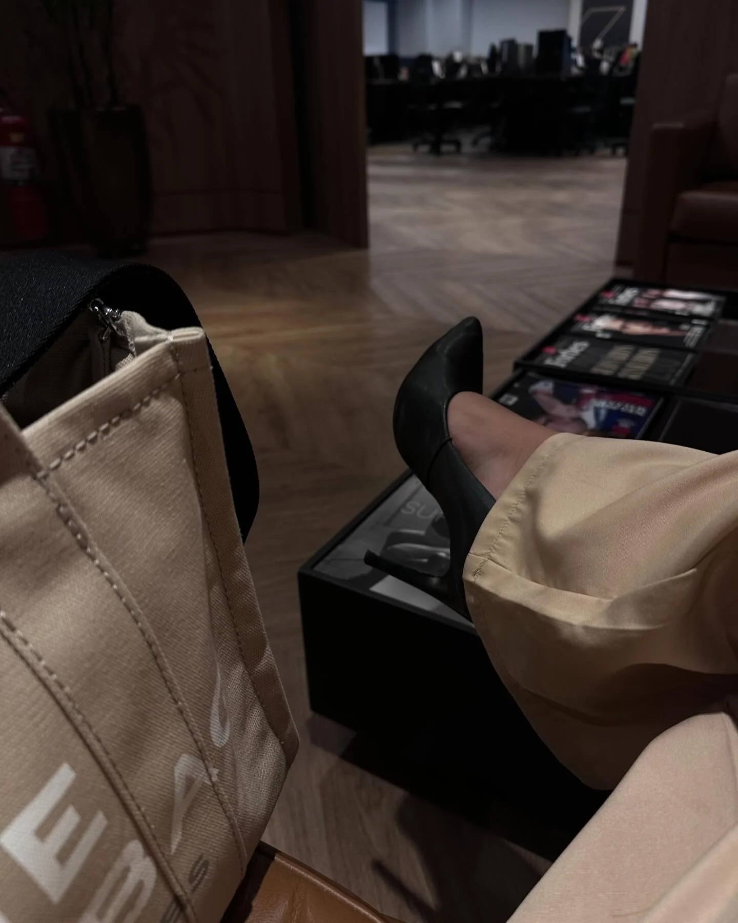 Person sitting with one leg crossed, wearing black shoes and beige pants, in a waiting area with magazines on the floor, a tan bag nearby, and a dark room in the background.