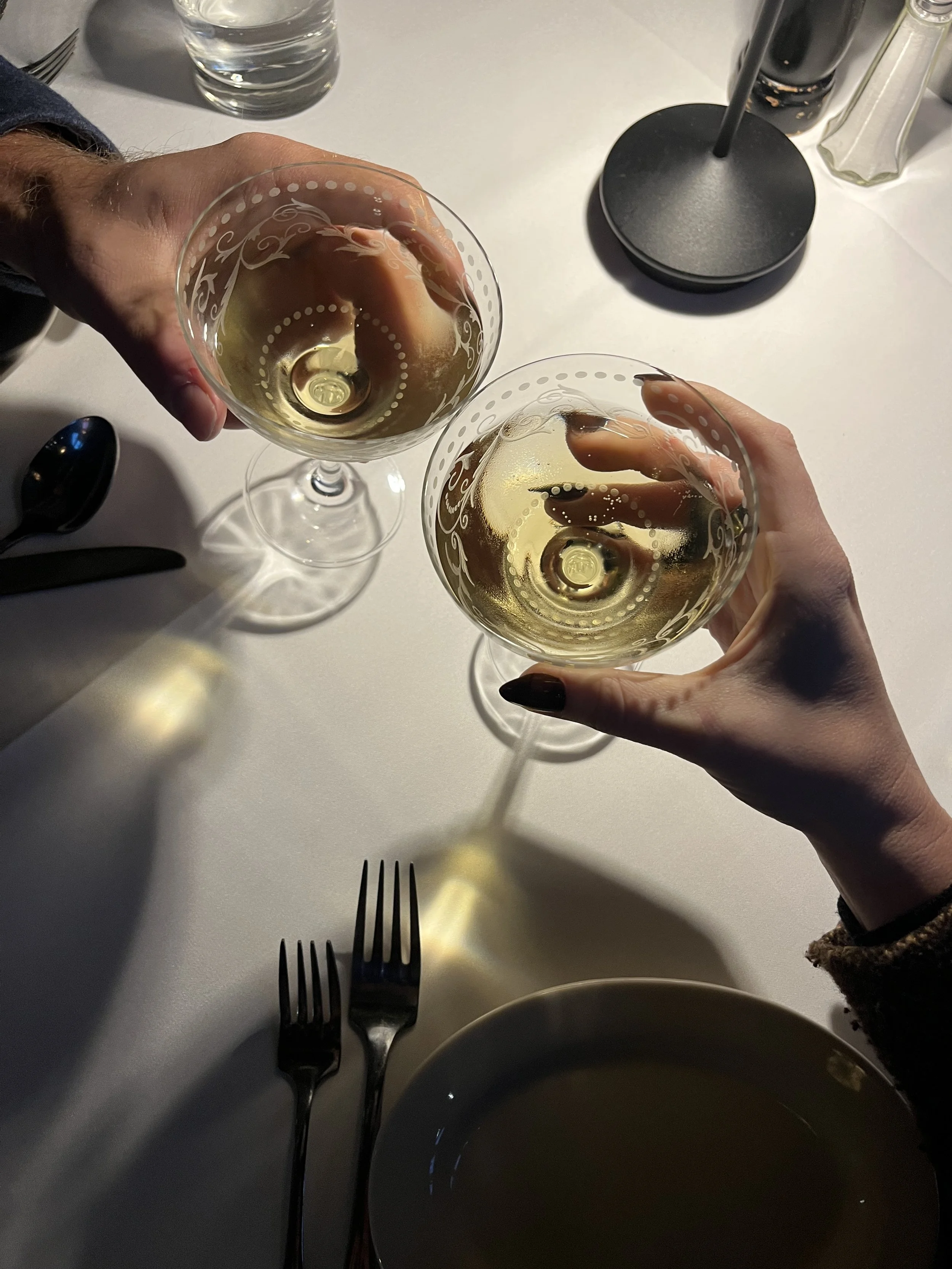 Two people clinking glasses of white wine at a dining table with two forks, a glass of water, a black plate, and a table lamp.