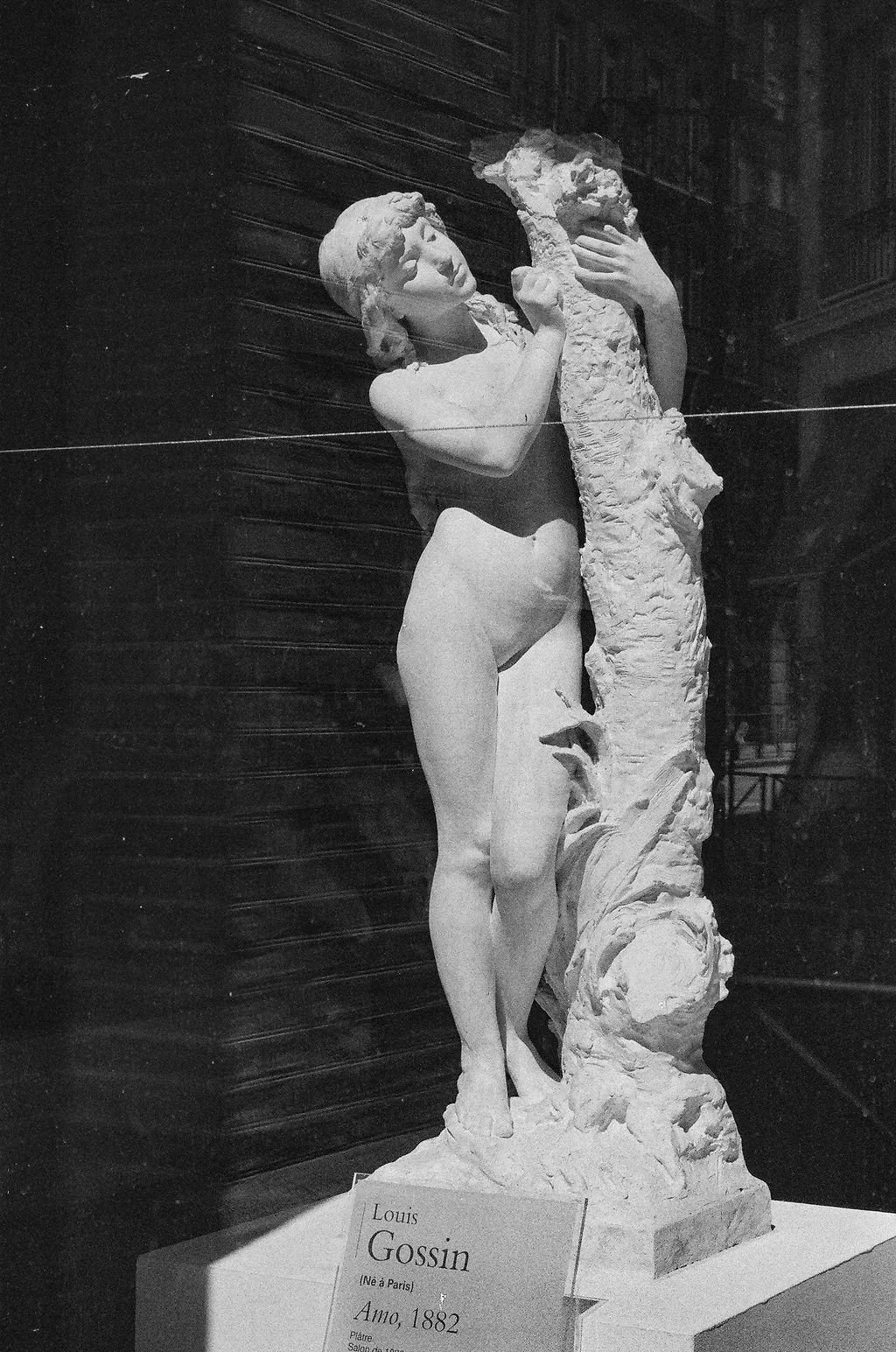 A sculpture of a woman holding a dog, displayed in an art gallery. The sculpture is reminiscent of classical art and is made of white material. There is a plaque below with the inscription "Louis Gossin, born in Paris, 1882, painter."