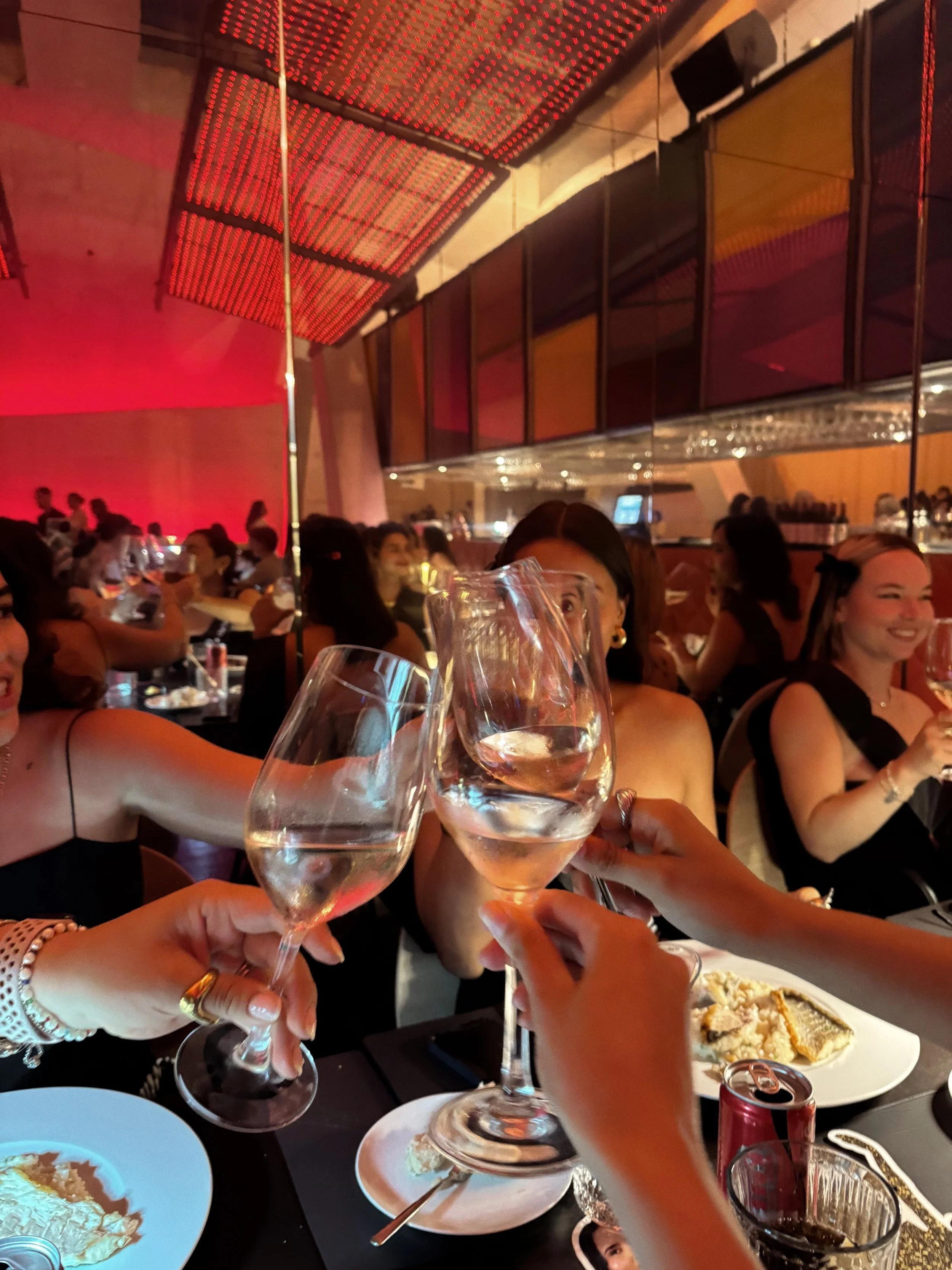 People at a restaurant table raising wine glasses for a toast, with plates of food and a lively, decorated interior in the background.