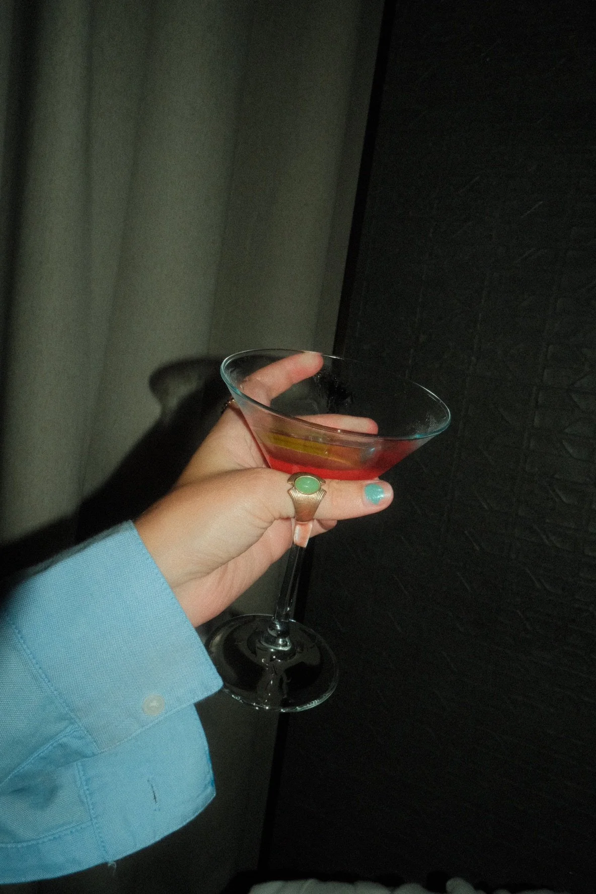 Person holding a martini glass with a pink cocktail, wearing a light blue long sleeve shirt and a ring with a green stone.
