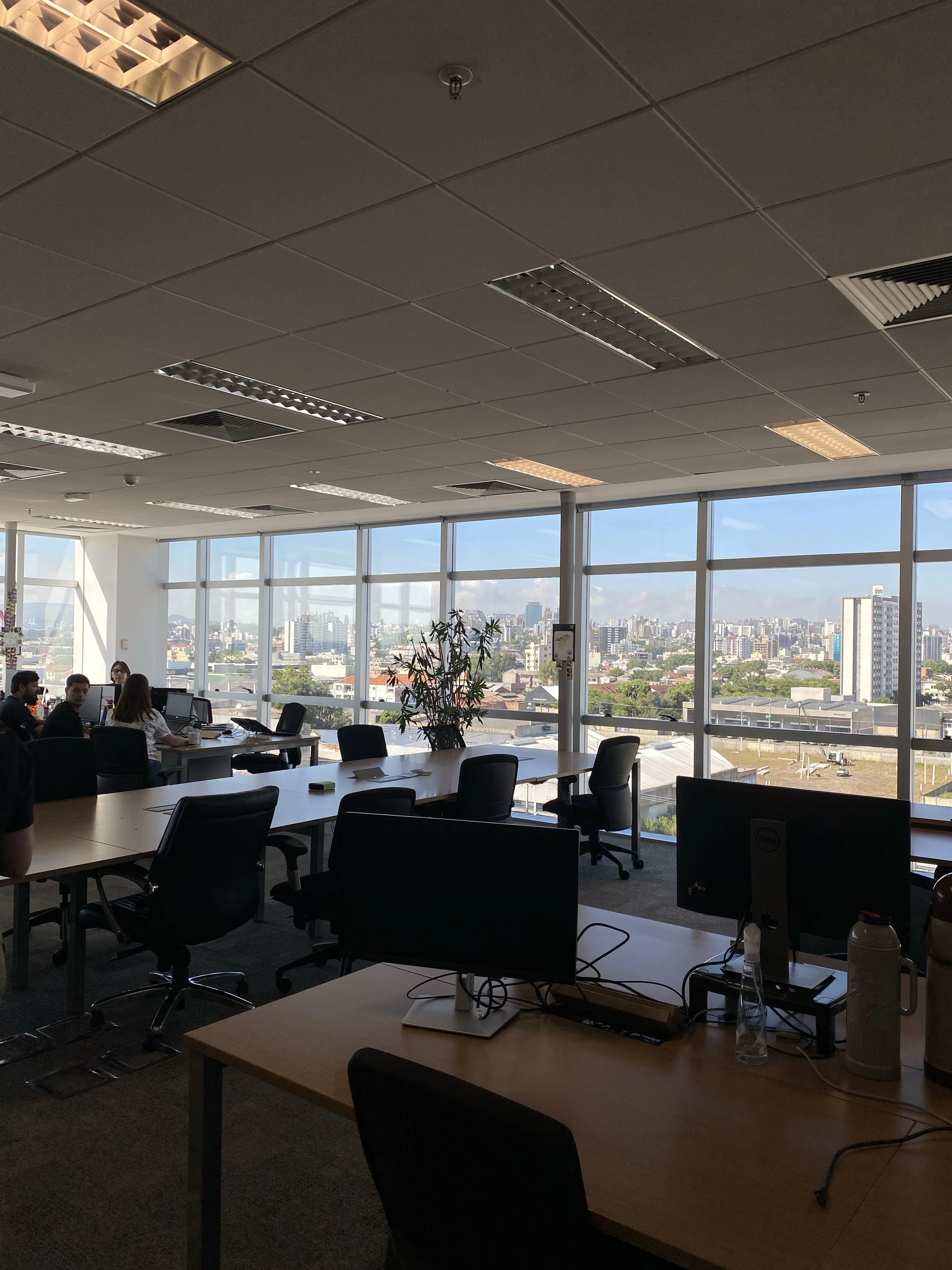 An office space with large windows showing a cityscape, several desks with computer monitors, and a group of people working.