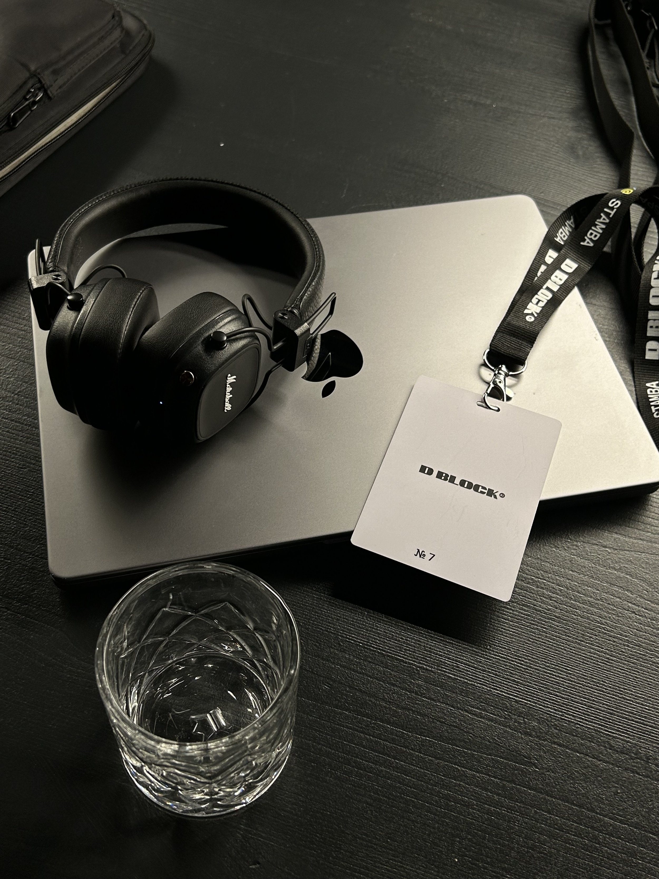 Black headphones resting on a closed silver laptop, with a glass of water in the foreground and various items in the background on a dark desk.