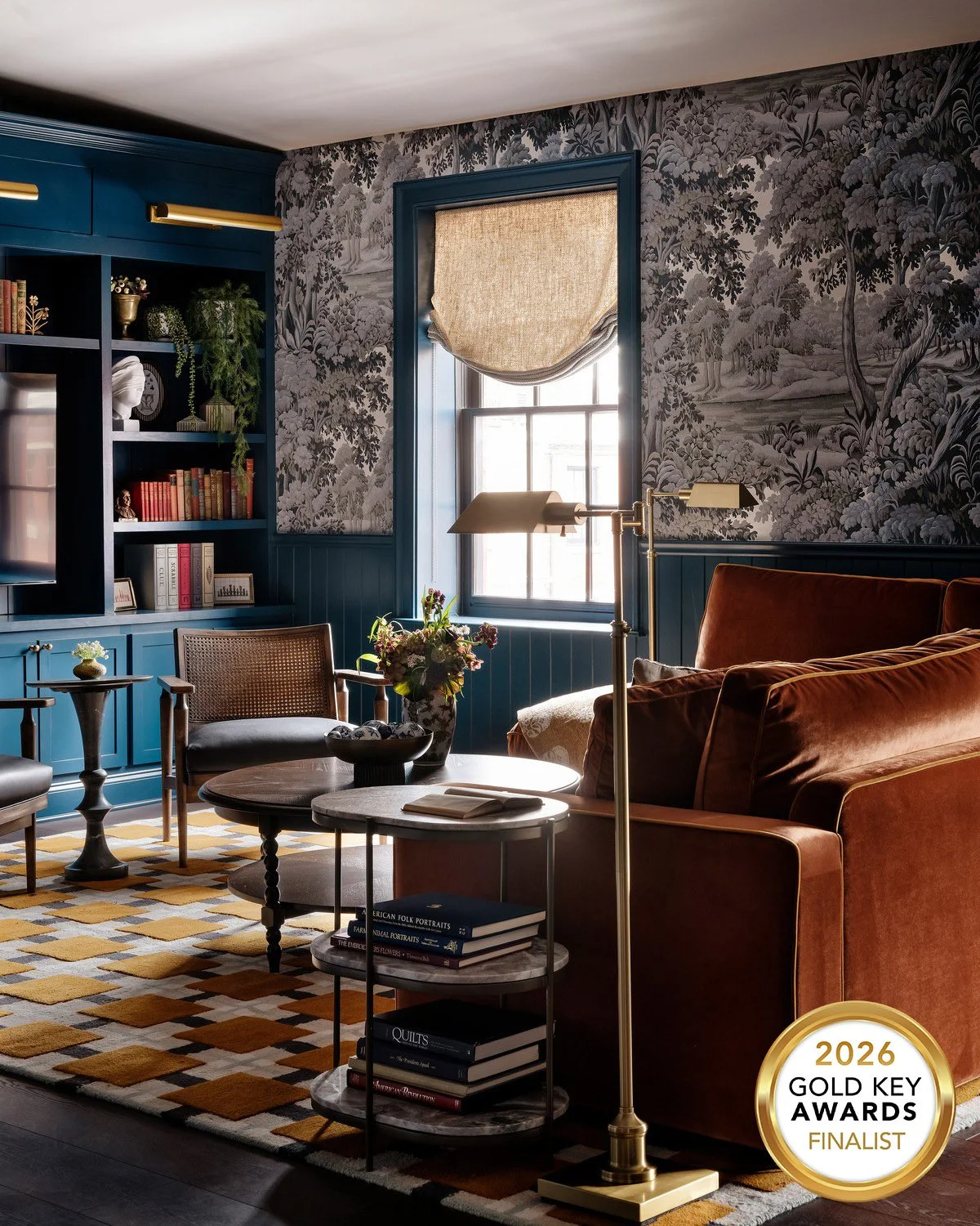 Envoy Suite living room at The Diplomat by Studio MaRō, with rust velvet sofa, navy built-in bookshelves, grisaille forest wallpaper and checkered rug. 2026 Gold Key Awards Finalist.