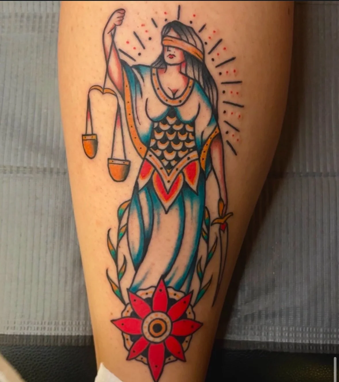 Tattoo of Lady Justice with scales, blindfold, sword, and a red flower design on a leg.