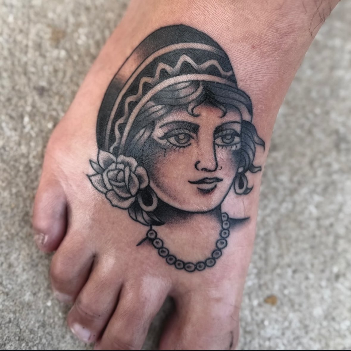 Tattoo of a woman's face with a retro style on a foot, featuring a rose and a pearl necklace.