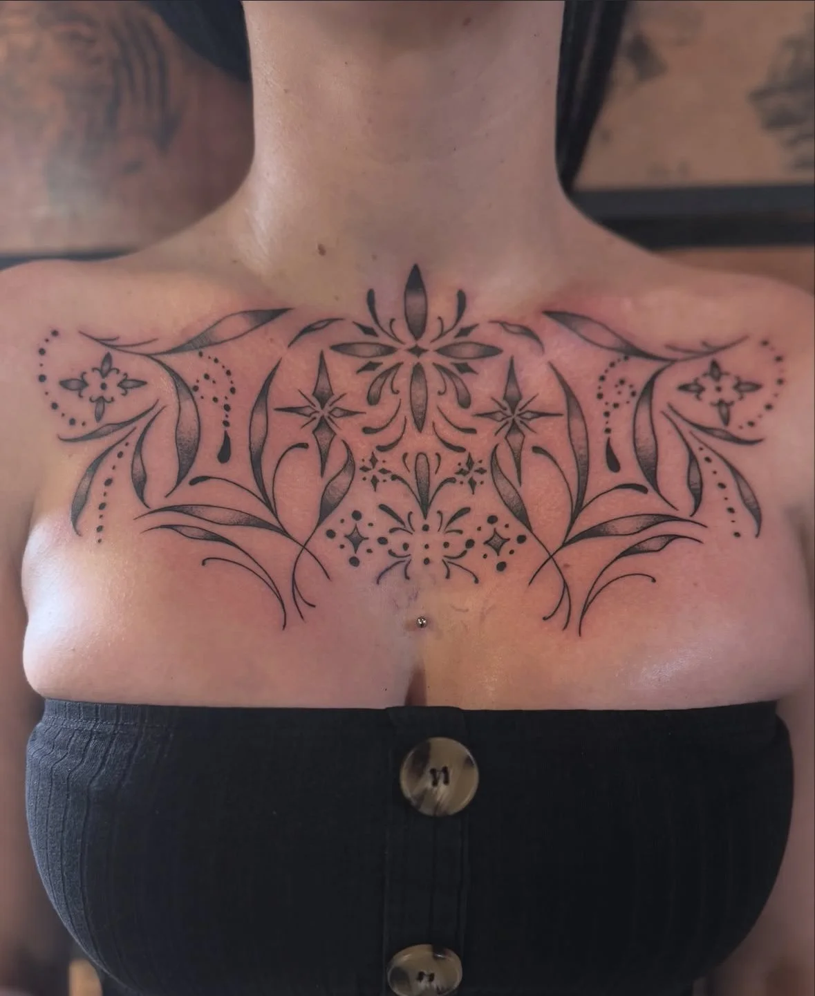 Intricate black ink chest tattoo with floral and geometric designs on a person wearing a black top with buttons.