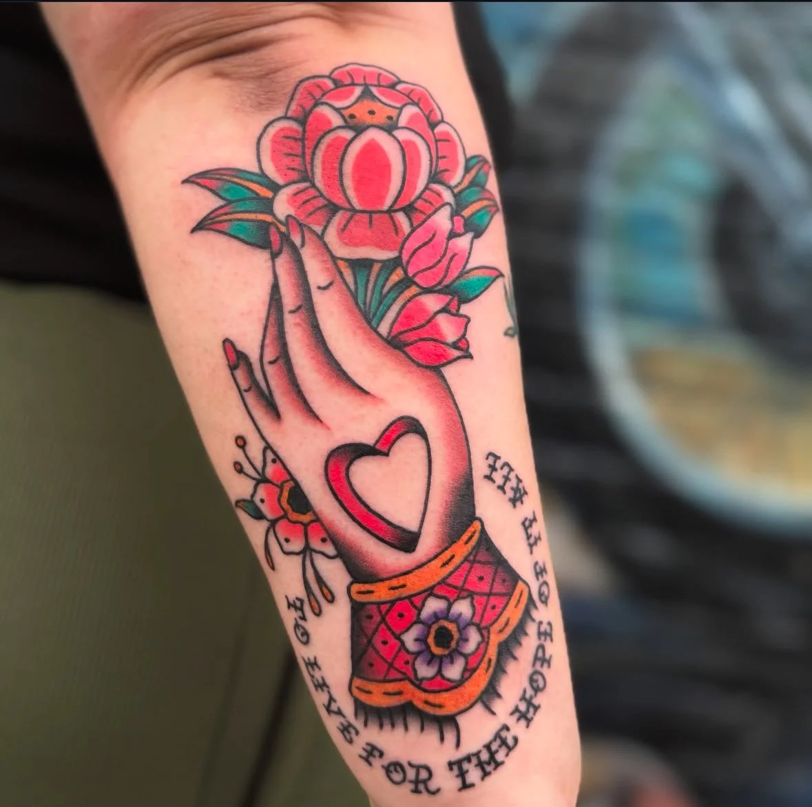 Tattoo of a hand holding a flower with heart symbol on the wrist and colorful floral designs on an arm.