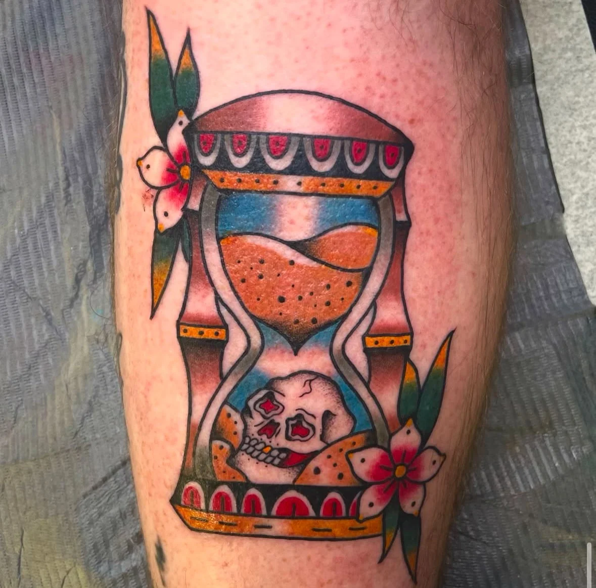 Tattoo of an hourglass with sand and skull, adorned with flowers and leaves.
