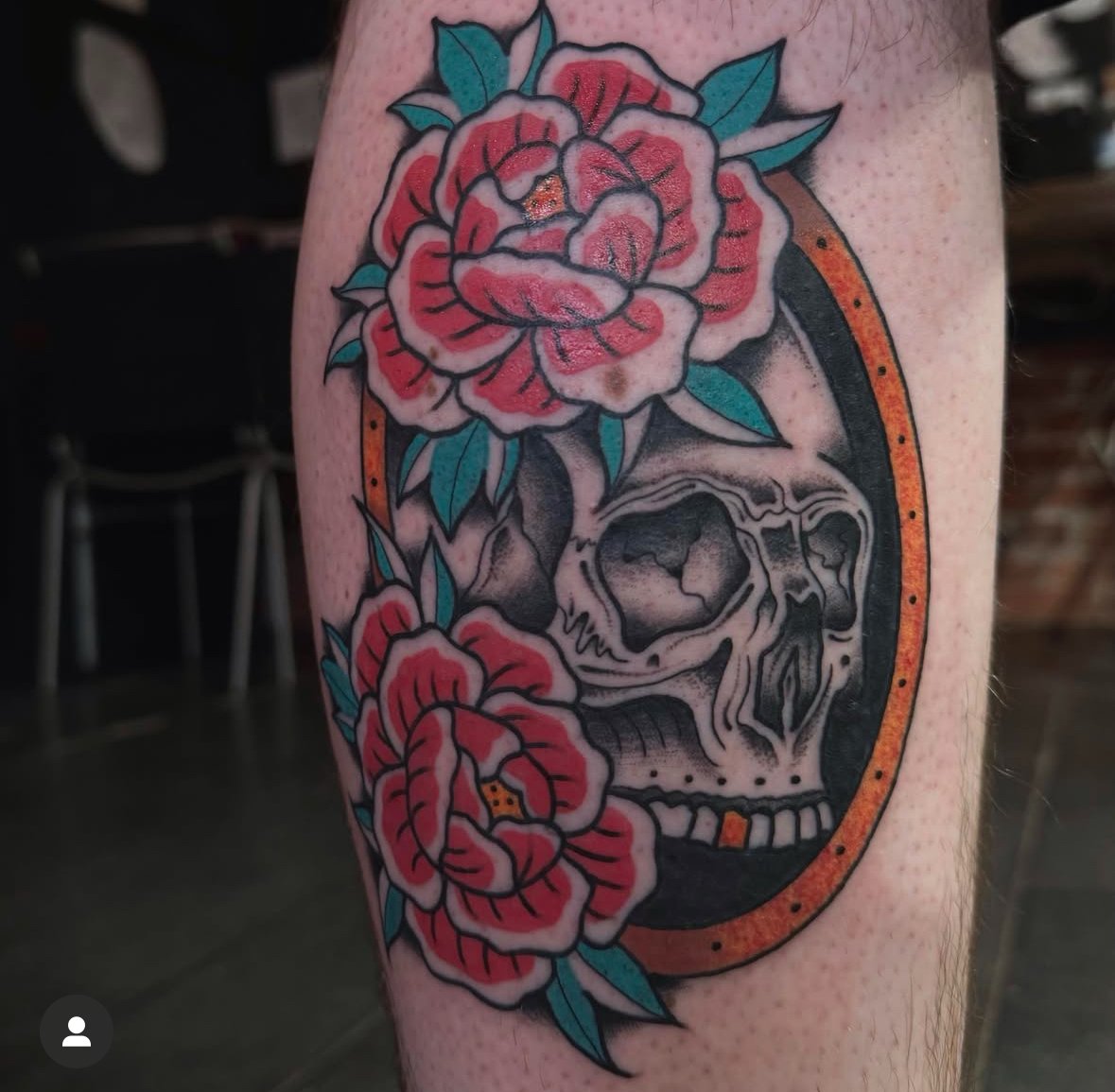 Tattoo of a skull surrounded by red flowers with green leaves on skin
