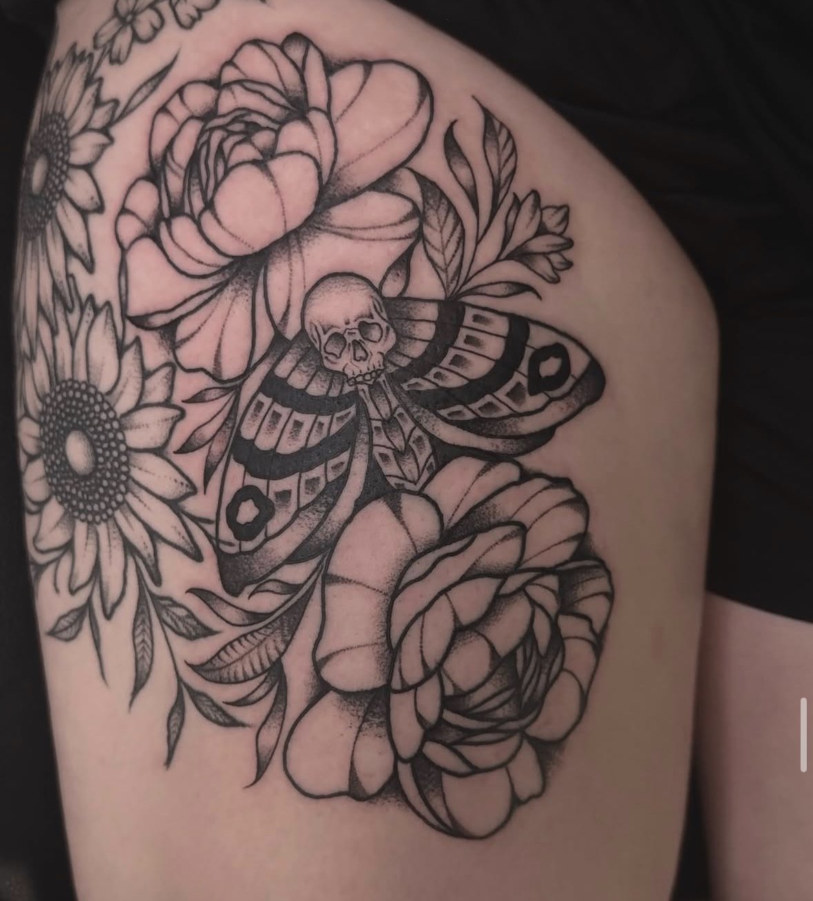 Tattoo of a moth with a skull on its back surrounded by roses and daisies on skin.