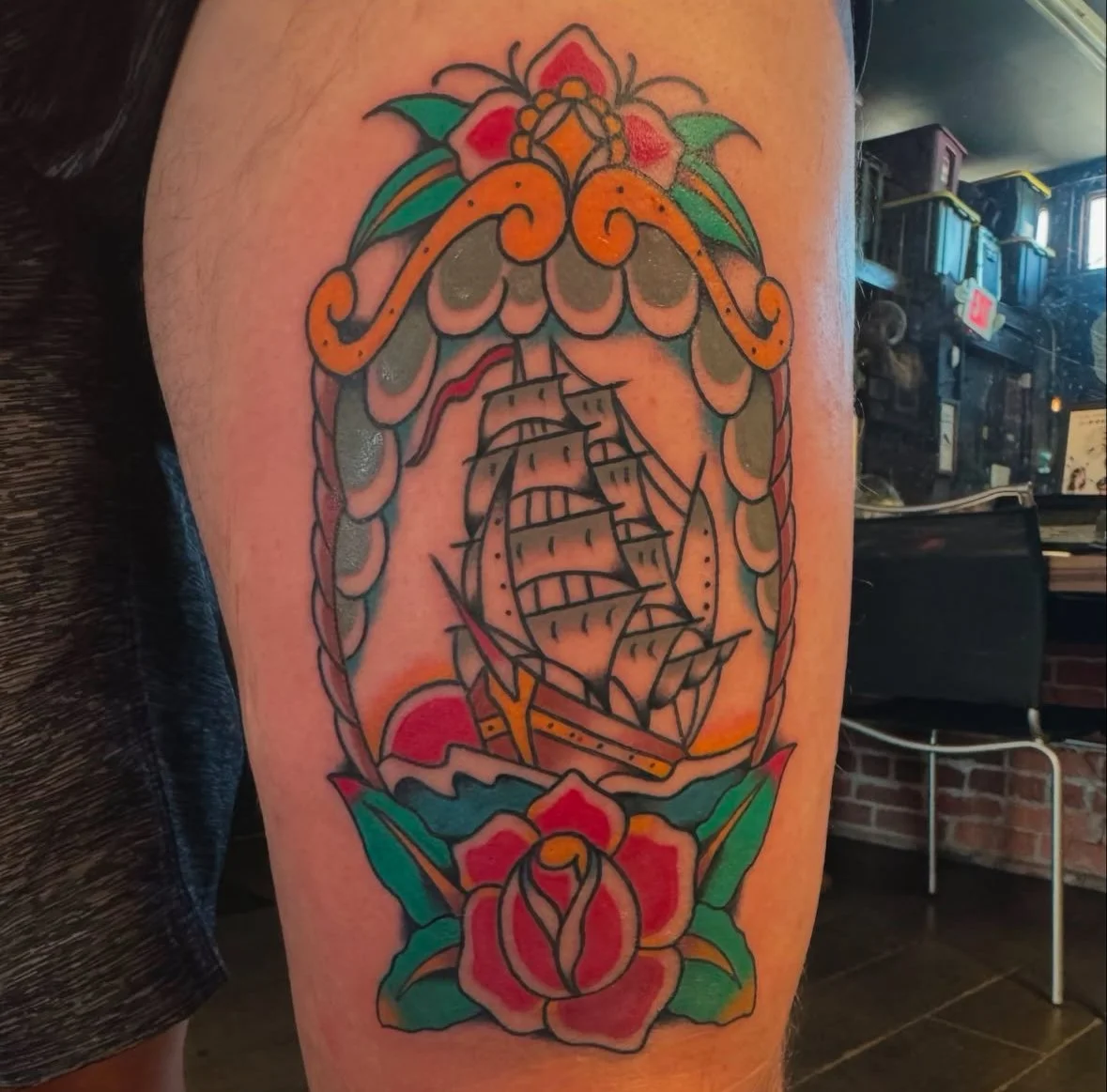 Tattoo of a traditional ship inside an ornate frame with flowers on skin.