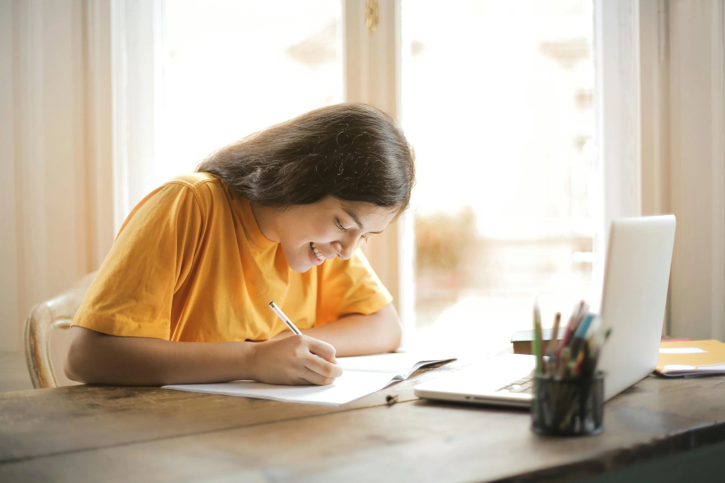 A student writing in a notebook with a laptop open in front of them