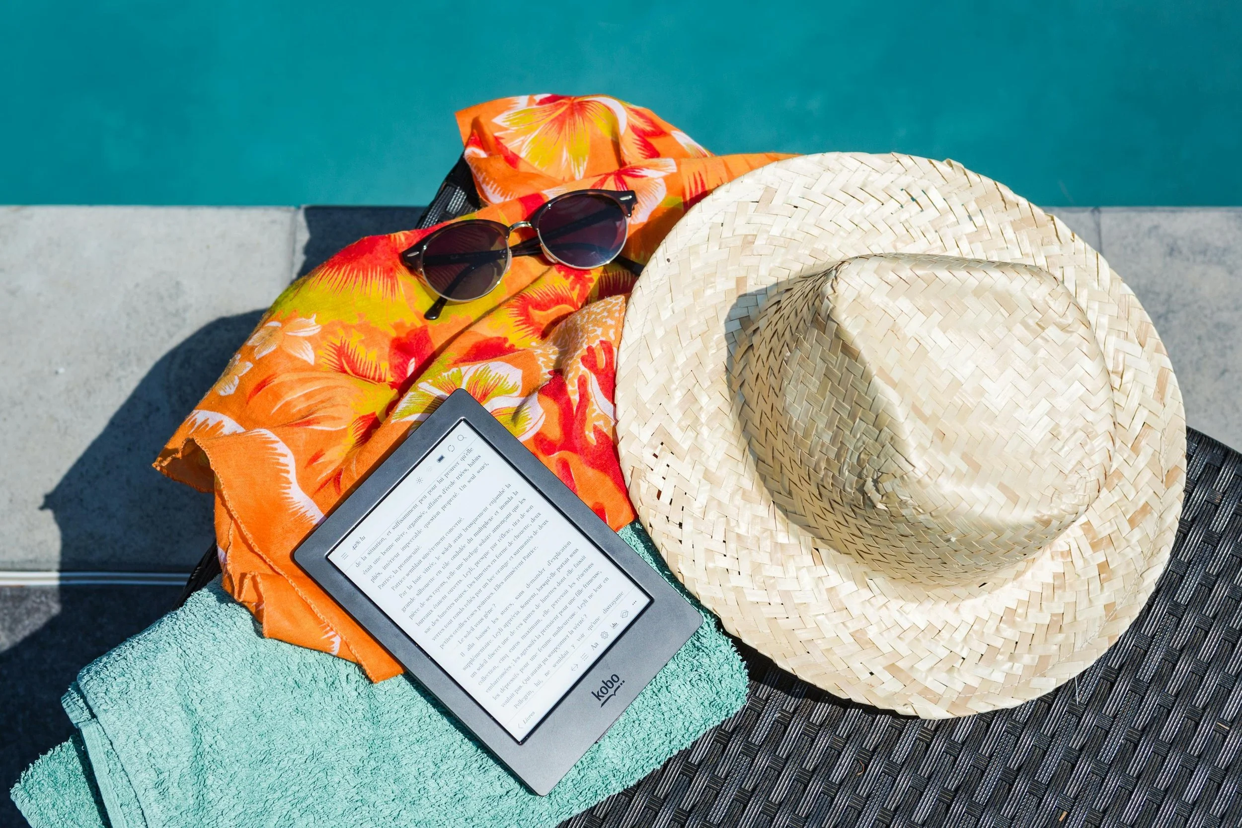 An e-reader sitting next to a hat and towel on a poolside chair