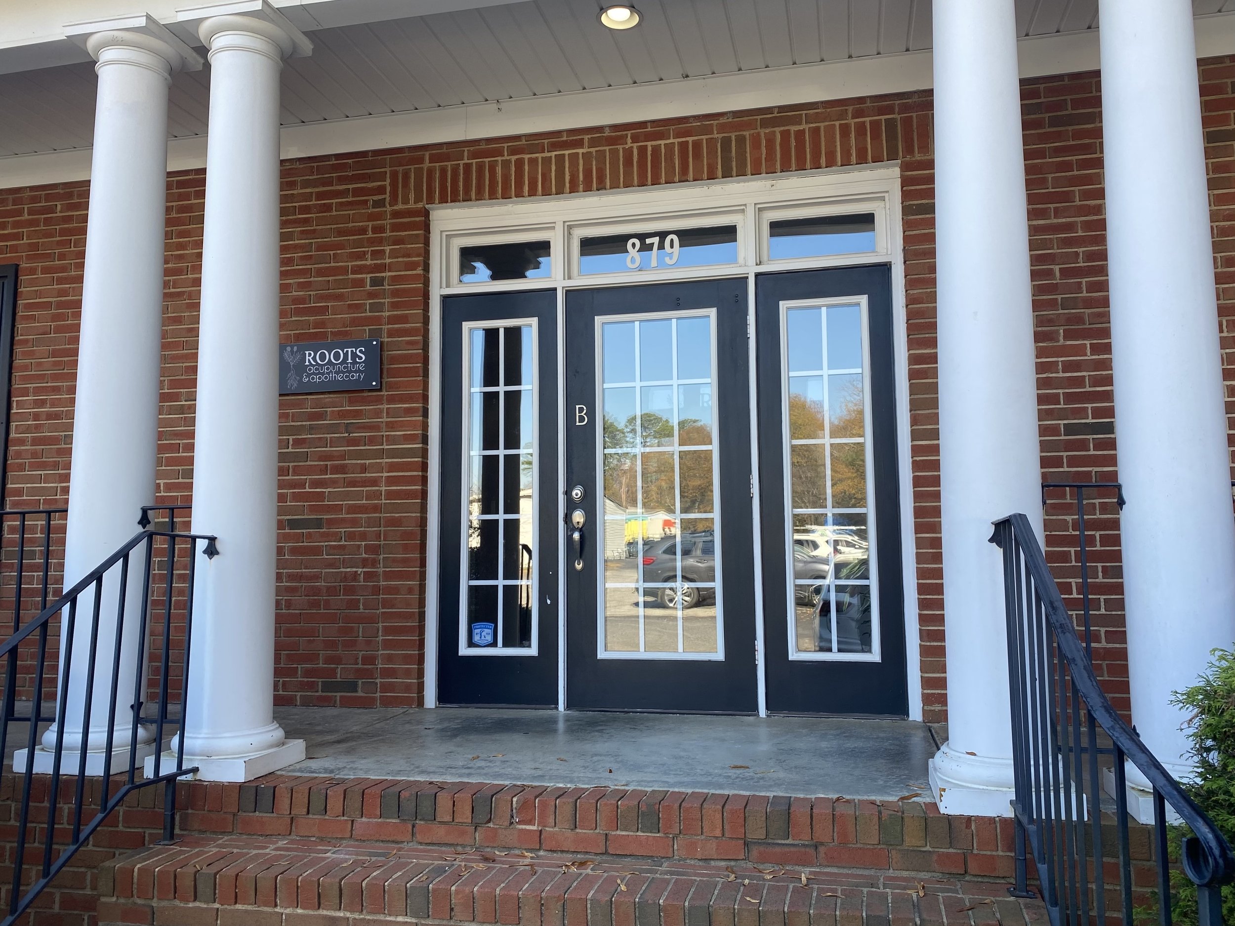The photo shows the entrance of a building with double glass doors, surrounded by a brick facade and white columns. The address 879 is above the door, and a sign reads 'ROOTS Acupuncture & Apothecary' to the left of the door.