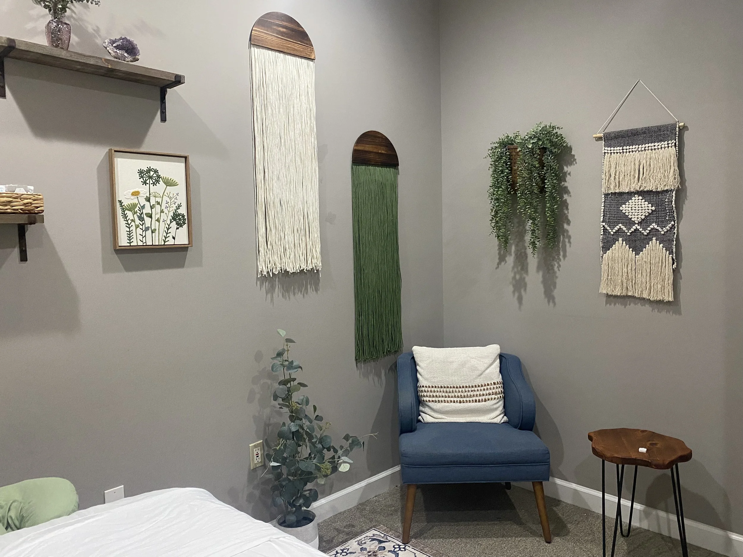 A corner of a room with gray walls decorated with plants, woven wall hangings, a blue armchair with a white pillow, a small wooden side table, and a massage table with white sheets and a green pillow.