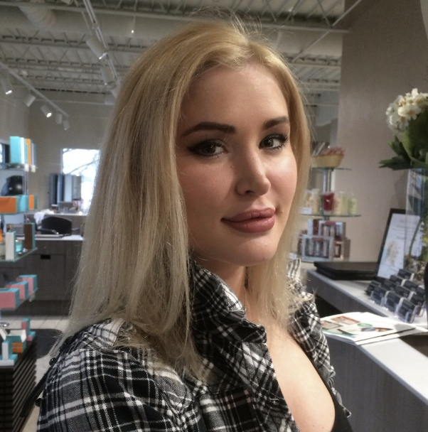 A woman with long blonde hair and makeup, wearing a black and white plaid shirt, standing inside a bookstore or gift shop with books, flowers, and framed photos in the background.